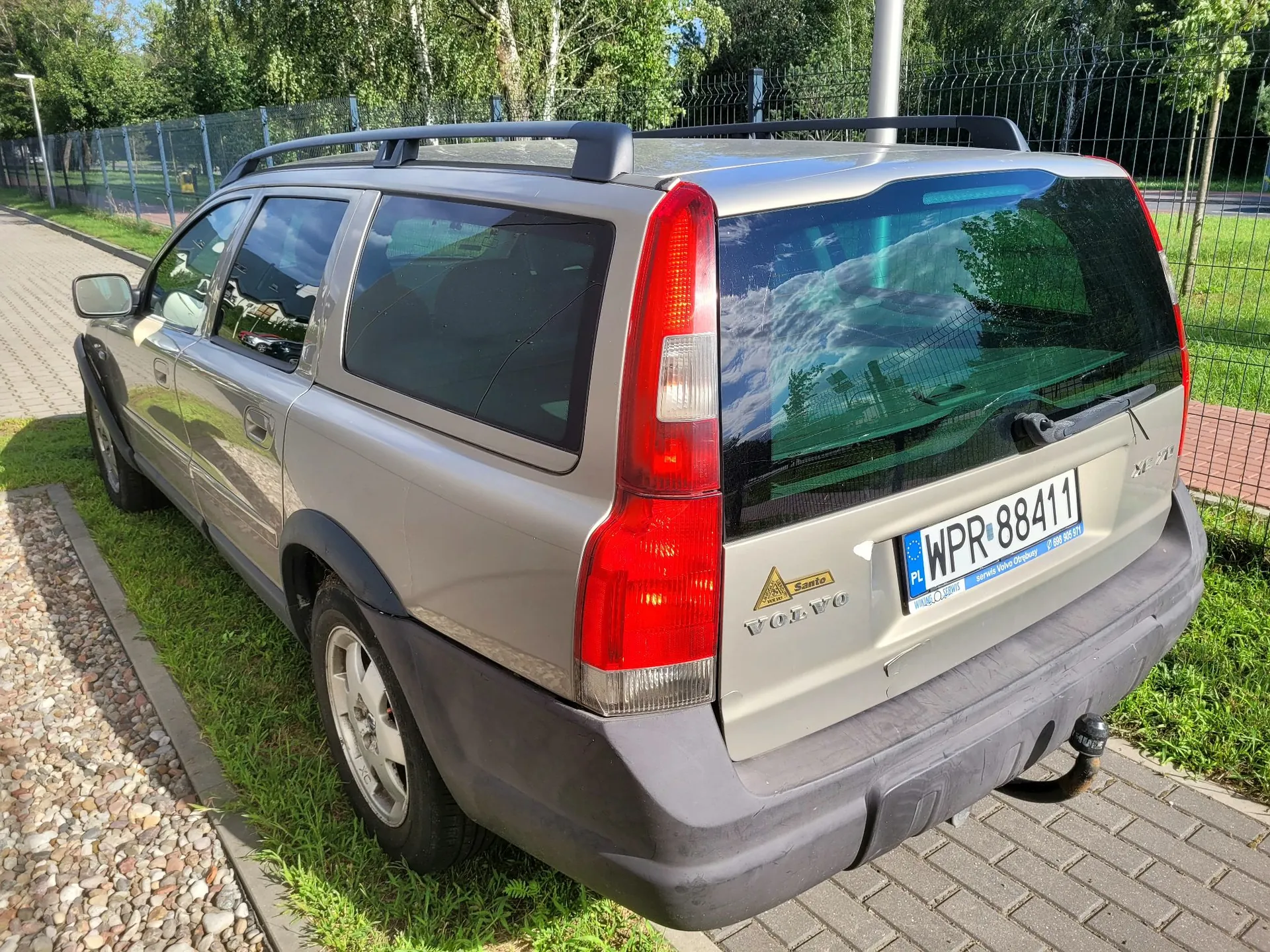 Srebrny samochód marki Volvo w nadwoziu kombi, zaparkowany na kostce brukowej obok trawnika i ogrodzenia z siatki. Widoczny jest tył pojazdu z polską tablicą rejestracyjną o numerze WPR 88411. Na klapie bagażnika znajdują się napisy 'VOLVO' oraz naklejka 'Santo'. Samochód posiada czarne relingi dachowe i ciemnoszare plastikowe osłony nadkoli oraz zderzak.