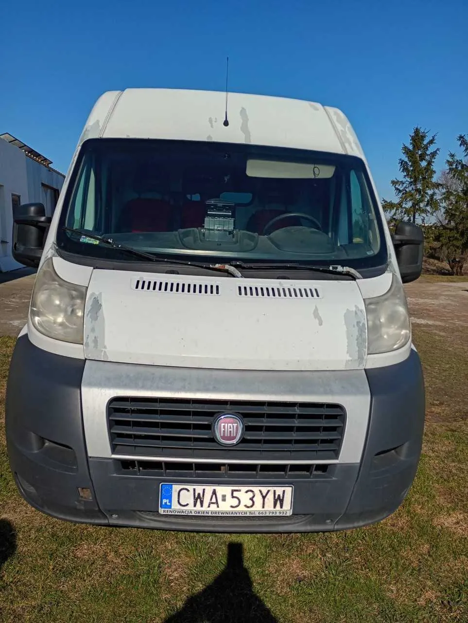 Biały samochód dostawczy marki Fiat z widocznym logo na atrapie chłodnicy. Na przedzie tablica rejestracyjna PL CWA-53YW oraz napis poniżej: RENOWACJA OKIEN DREWNIANYCH tel. 663 793 932. Samochód ma uszkodzenia lakieru na masce. W tle budynek po lewej stronie, drzewa po prawej oraz niebieskie niebo. Cień osoby fotografującej widoczny na trawie pod samochodem. Wnętrze pojazdu z czerwonymi siedzeniami i kierownicą.