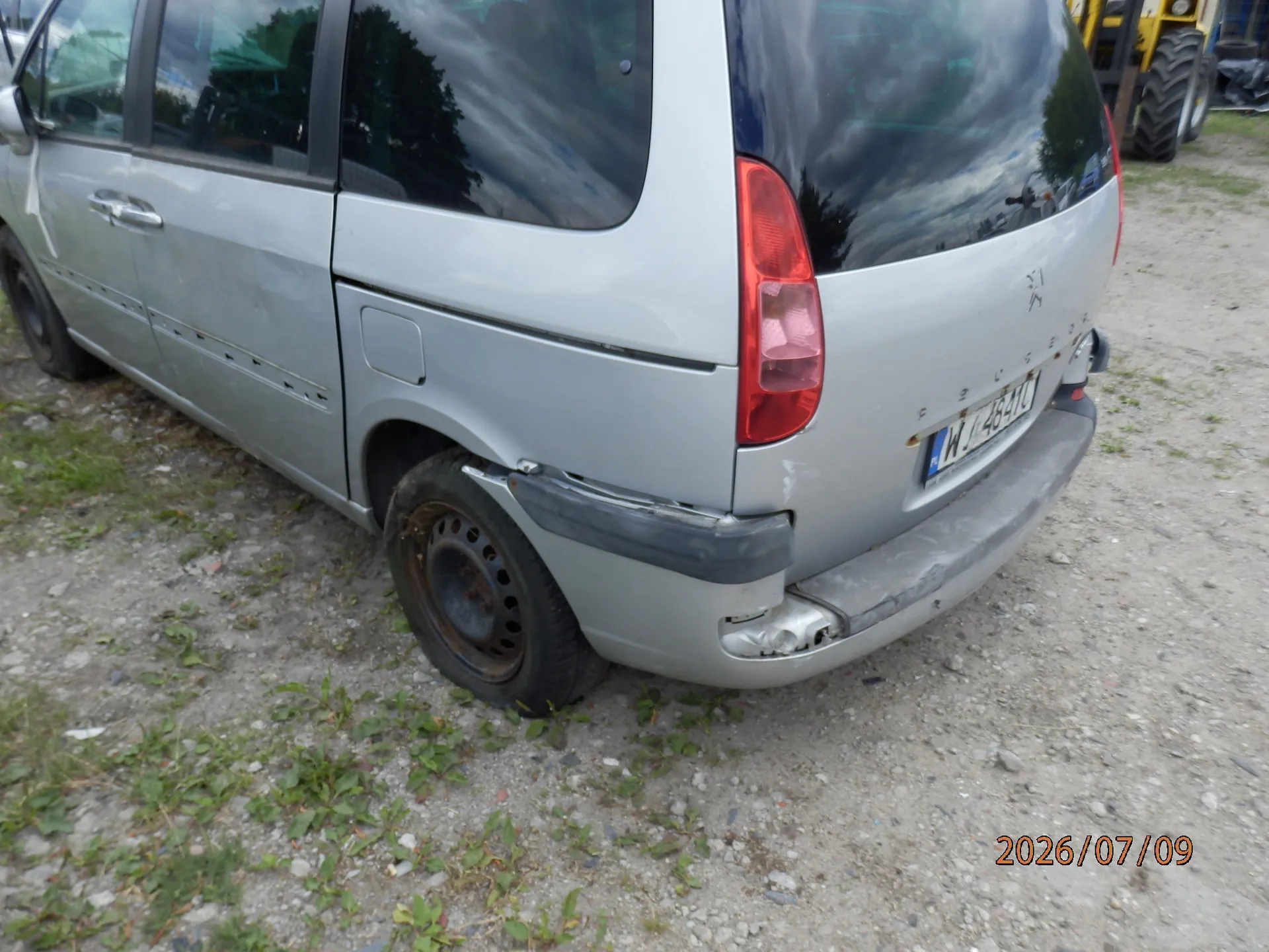 Srebrny samochód marki Peugeot typu minivan, widoczny z tyłu i z boku. Pojazd posiada uszkodzenia w postaci wgnieceń i zarysowań na tylnym zderzaku oraz na lewym tylnym błotniku. Widoczna jest tablica rejestracyjna o numerze WY 464YC. Samochód stoi na żwirowym podłożu. W prawym dolnym rogu zdjęcia znajduje się data 2026/07/09.