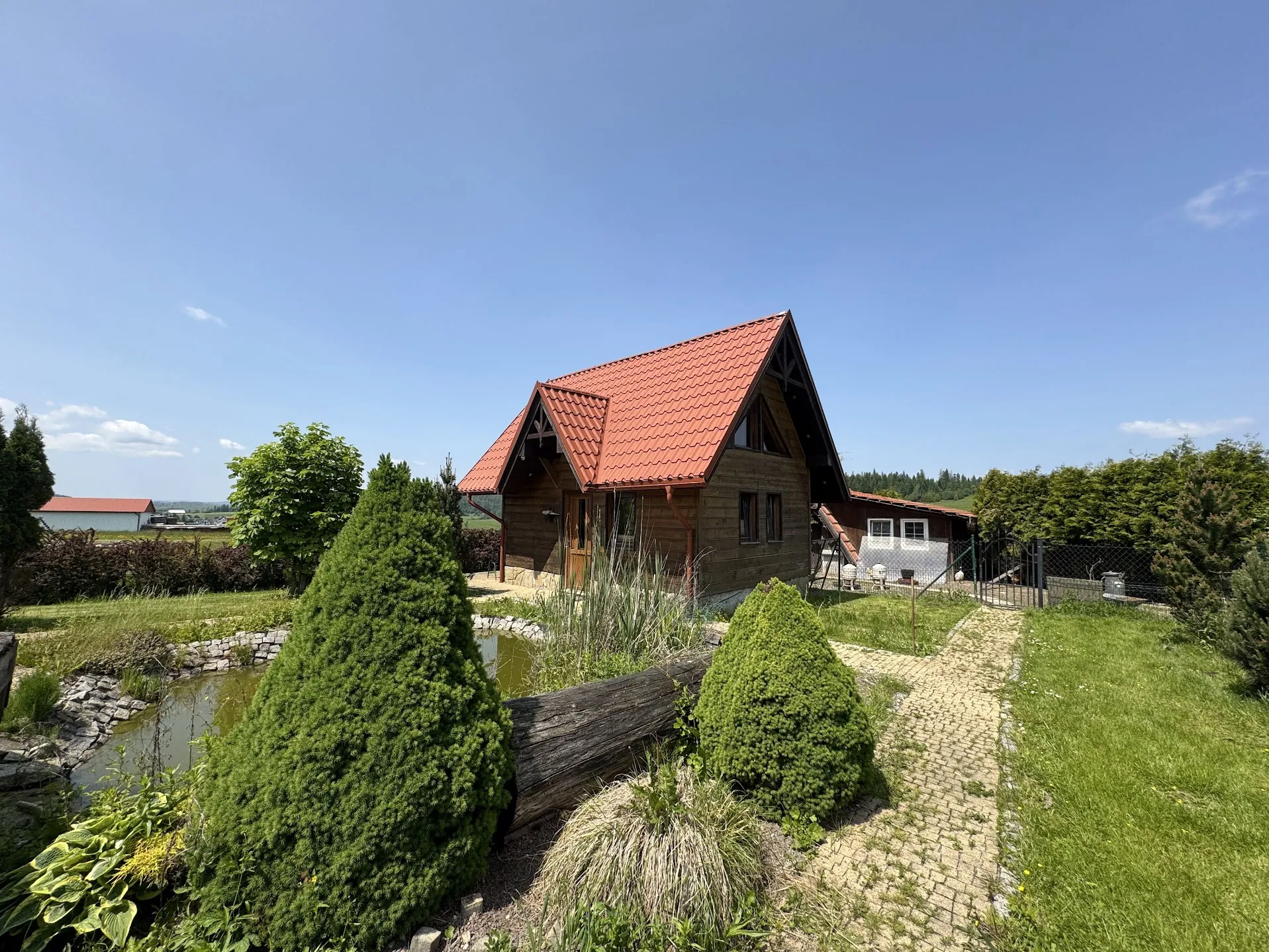 Drewniany dom z czerwoną dachówką, otoczony zielonym ogrodem z trawnikiem, krzewami, stawem wyłożonym kamieniami oraz ścieżką z cegieł. W tle widoczne są inne budynki, drzewa i jasne niebo z kilkoma chmurami. Dom ma brązowe ściany, okna i drzwi, a obok znajduje się ogrodzenie z siatki.