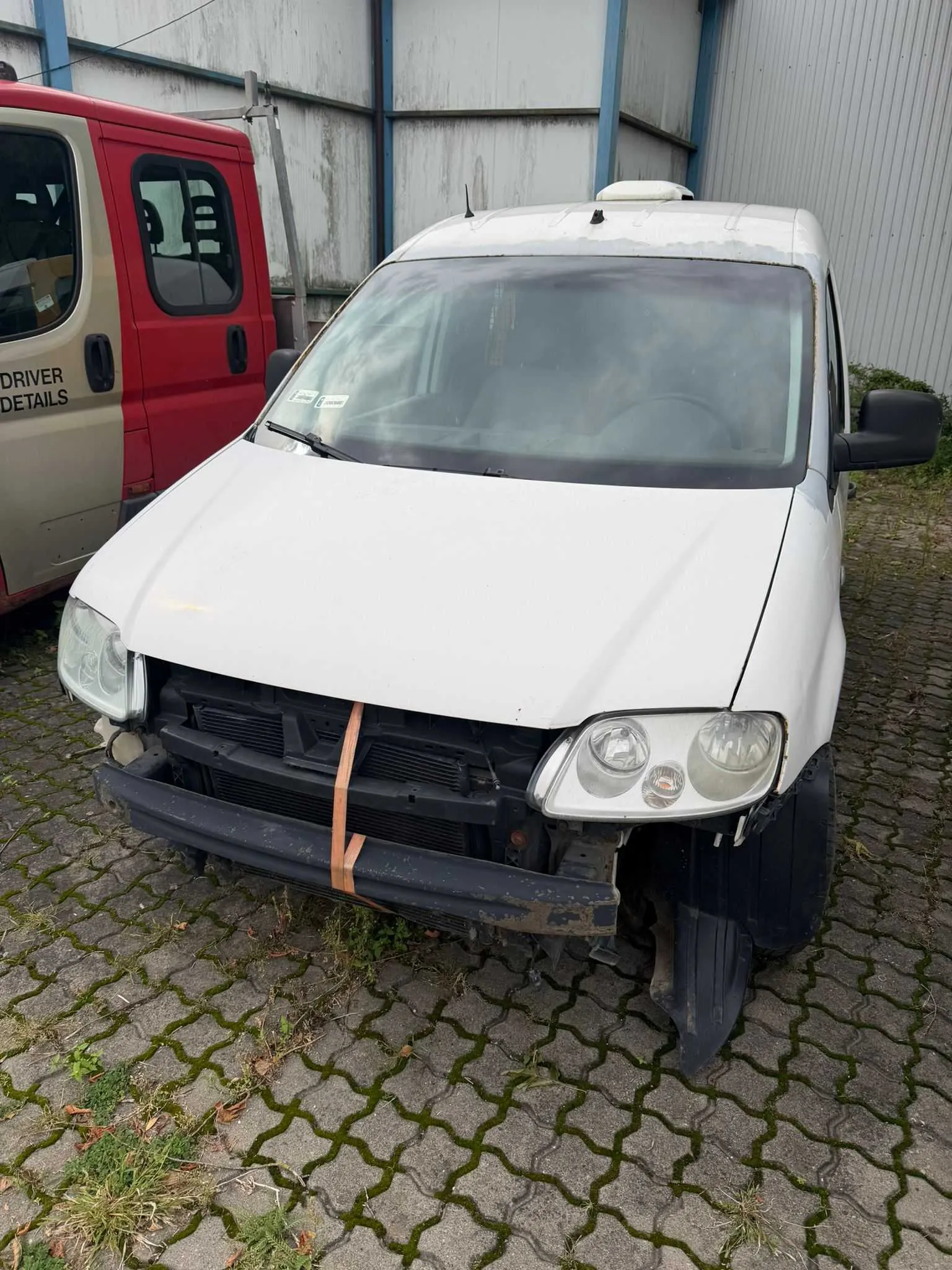 Biały samochód dostawczy z uszkodzonym przodem, zaparkowany na kostce brukowej. Brakuje przedniego zderzaka, a grill jest przymocowany do reszty nadwozia za pomocą pomarańczowej taśmy. Po lewej stronie widoczny jest fragment czerwonego vana z napisem 'DRIVER DETAILS'.
