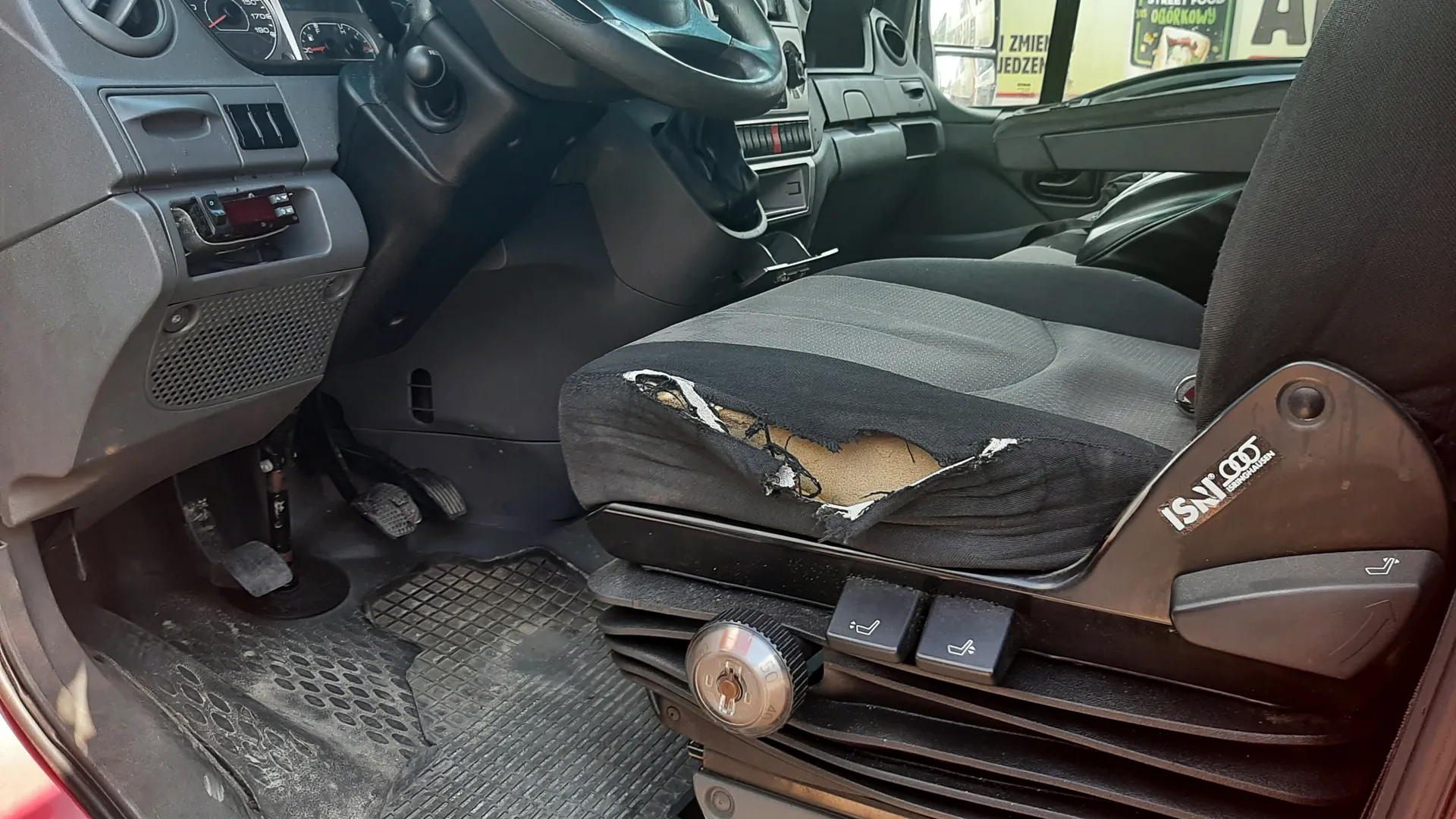 Wnętrze pojazdu z widocznym uszkodzonym siedzeniem kierowcy, na którym widnieje napis ISRI 900. Widoczne są kierownica, deska rozdzielcza z licznikami, pedały oraz brudne dywaniki. Przez okno widać fragment ulicy z napisami ZMIEN JEDZENIE i OGORKOWY na szybie innego pojazdu lub budynku. Siedzenie ma rozdarte pokrycie, odsłaniając piankę.