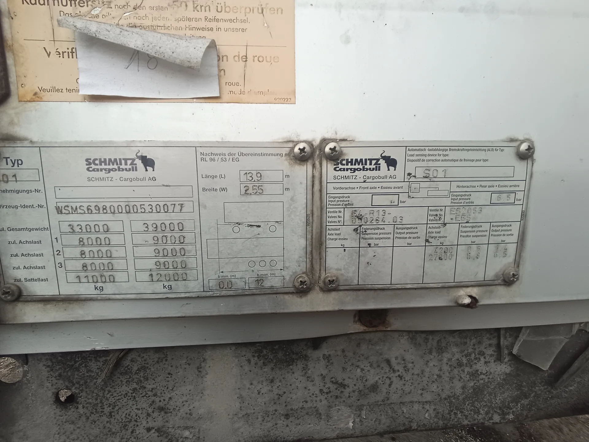Dwa metalowe tablice identyfikacyjne firmy SCHMITZ Cargobull AG zamocowane na białej powierzchni. Lewa tablica zawiera dane: Typ 01, numer identyfikacyjny pojazdu WSMS6980000530077, wagi (33000 kg, 39000 kg), dopuszczalne obciążenia osi (8000/9000 kg dla osi 1-3), długość 13,9 m, szerokość 2,55 m. Prawa tablica zawiera parametry techniczne: numer S01, dane zaworów (np. 090264.03), ciśnienia wejściowe/wyjściowe (6,5 bar), obciążenia osi (4200 kg, 27000 kg). Nad tablicami widoczny jest częściowo zniszczony papier z tekstem w języku niemieckim (m.in. 'nach den ersten 50 km überprüfen') i francuskim (m.in. 'Vérif', 'roue'). Tło: biała powierzchnia z widocznymi śladami korozji i śrubami.