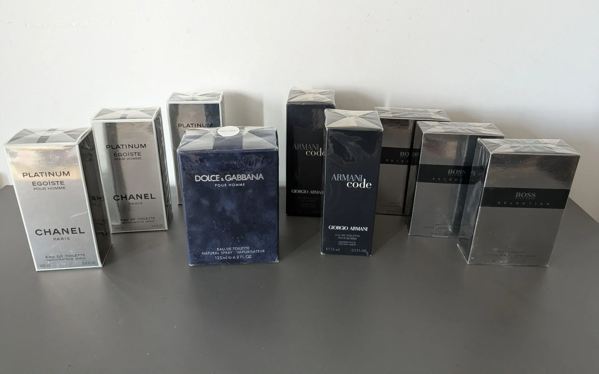 Kolekcja dziesięciu kartonowych opakowań męskich perfum ustawionych w rzędzie na szarej powierzchni na tle białej ściany. Od lewej strony znajdują się trzy opakowania Chanel Platinum Égoïste, następnie dwa opakowania Dolce & Gabbana Pour Homme, dwa opakowania Armani Code oraz trzy opakowania Boss Selection.