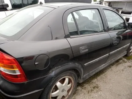 Czarny samochód osobowy typu sedan zaparkowany na żwirowym podłożu. Widok z tyłu i lewej strony. Samochód posiada czerwone tylne światła, srebrne felgi pięcioramiennych i widoczne zabrudzenia na dolnej części nadwozia. W tle częściowo widoczne są inne pojazdy oraz fragmenty budynków.