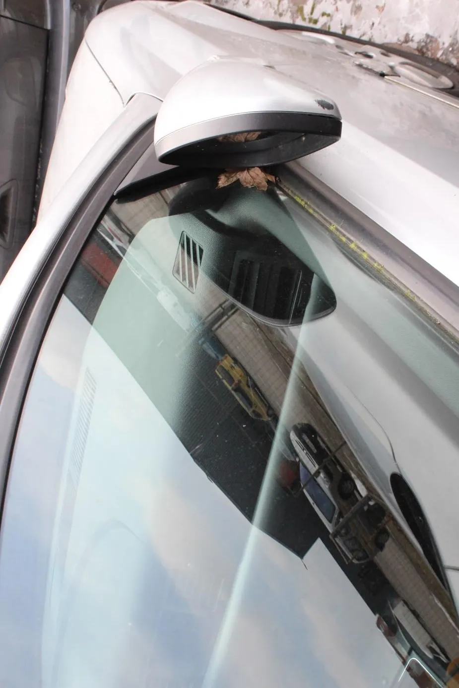 Boczny widok srebrnego samochodu z widocznym lusterkiem bocznym i przednią szybą. W odbiciu na szybie widać inne pojazdy: żółty van i biały samochód, a także ceglany budynek i niebo. Przy lusterku bocznym znajduje się suchy liść. Na ziemi widoczne są plamy śniegu.