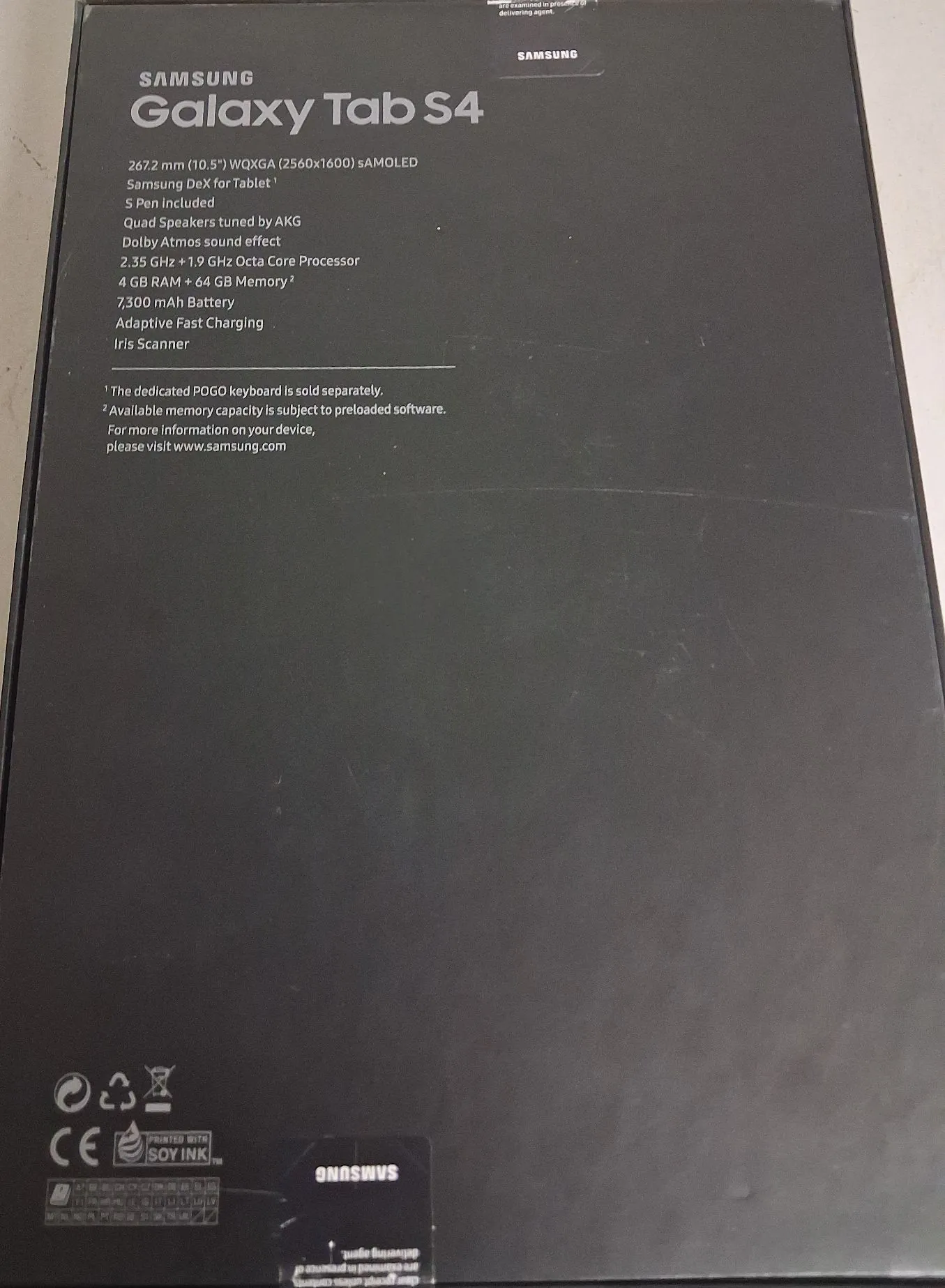 Zdjęcie tylnej części czarnego opakowania tabletu Samsung Galaxy Tab S4. Na górze znajduje się logo SAMSUNG oraz nazwa modelu Galaxy Tab S4. Poniżej wypisana jest specyfikacja techniczna: ekran 267,2 mm (10,5") WQXGA (2560x1600) sAMOLED, Samsung DeX for Tablet, S Pen w zestawie, głośniki Quad Speakers dostrojone przez AKG, Dolby Atmos, procesor 2.35 GHz + 1.9 GHz Octa Core, 4 GB RAM + 64 GB pamięci, bateria 7,300 mAh, Adaptive Fast Charging oraz Iris Scanner. Na dole znajdują się przypisy dotyczące klawiatury POGO i pamięci, adres strony www.samsung.com oraz symbole certyfikacji, w tym znak CE i informacja PRINTED WITH SOY INK. Na samym dole widoczne jest odwrócone logo SAMSUNG.