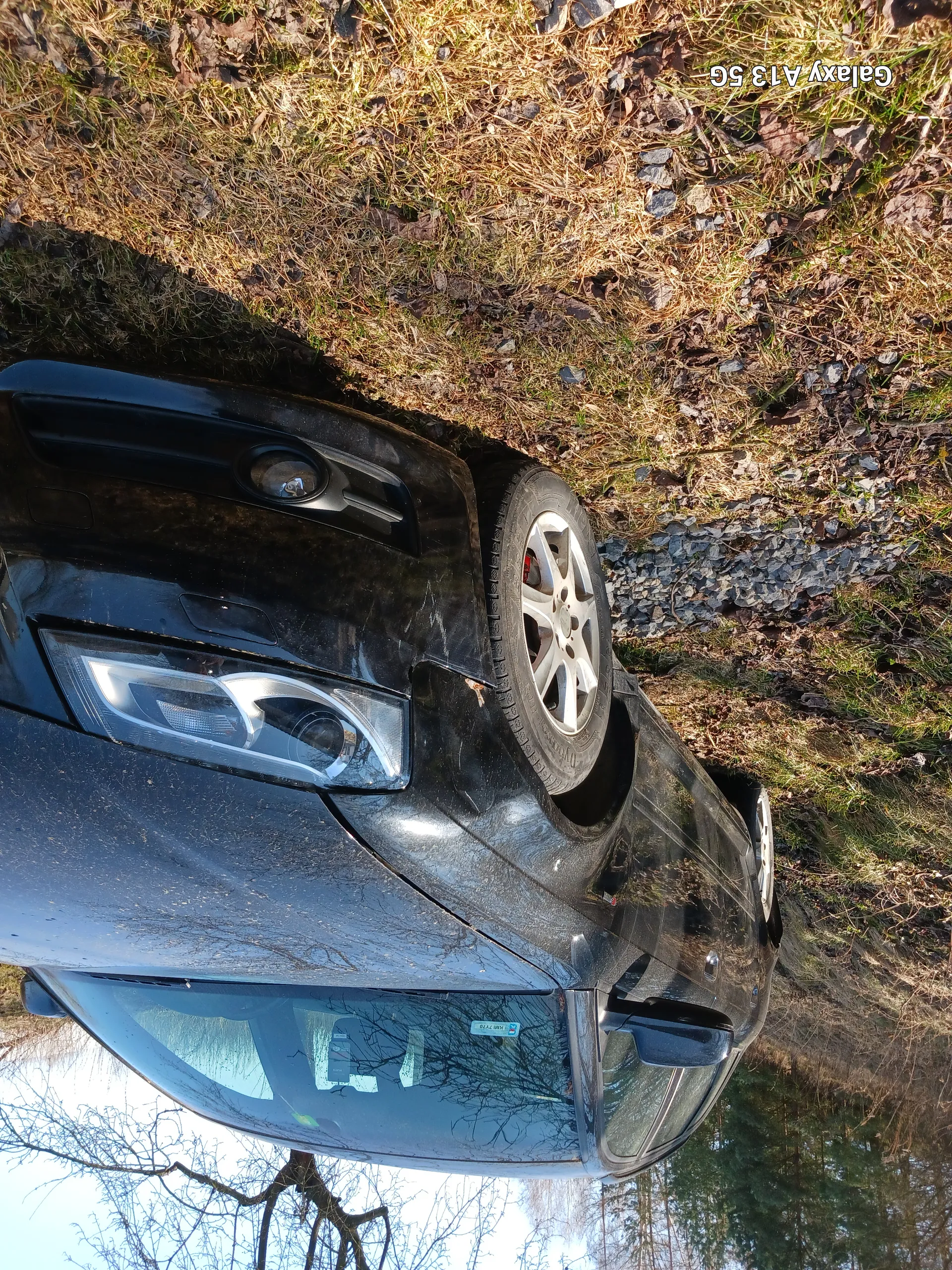 Czarny samochód zaparkowany na trawie i żwirze. Widoczna przód z reflektorem, błotnikiem i kołem z srebrną felgą. Samochód jest zabrudzony. W tle drzewa bez liści, zielone drzewa i niebo. Po prawej stronie obrazu widoczny tekst 'Galaxy A13 5G'.