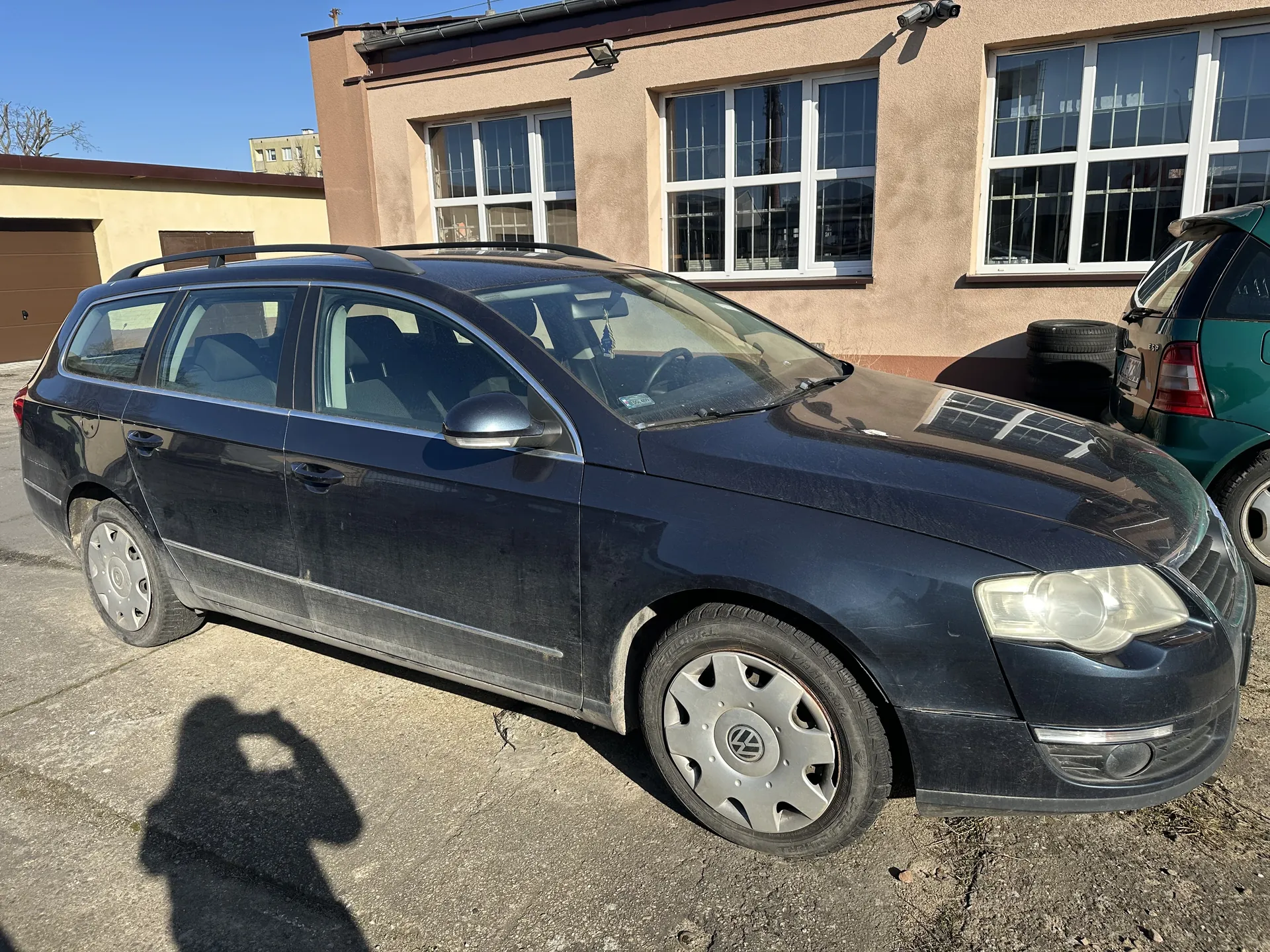 Ciemnoniebieski samochód osobowy typu kombi marki Volkswagen zaparkowany na betonowym placu. Pojazd posiada dachówkę, srebrne felgi z logo VW oraz zanieczyszczoną karoserię. W tle widoczny jest beżowy budynek z białymi oknami, stos opon oraz zielony samochód. Na asfalcie znajduje się cień osoby robiącej zdjęcie. Niebo jest bezchmurne i błękitne.