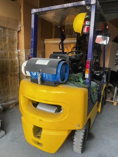 Żółty wózek widłowy w magazynie. Na przedniej części widoczny niebieski zbiornik gazowy, żółty kask i gaśnica. W tle stosy kartonów owiniętych folią. Wózek posiada kierownicę, siedzenie oraz lusterko boczne.