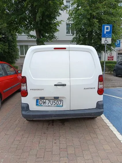 Tylny widok białego samochodu dostawczego marki Peugeot Partner zaparkowanego na kostce brukowej. Pojazd ma numer rejestracyjny DW 7U507. Po lewej stronie widoczny jest fragment czerwonego samochodu osobowego. W tle znajduje się biały budynek z oknami, zielone drzewa oraz niebieski znak parkingowy z symbolem osoby z niepełnosprawnością.