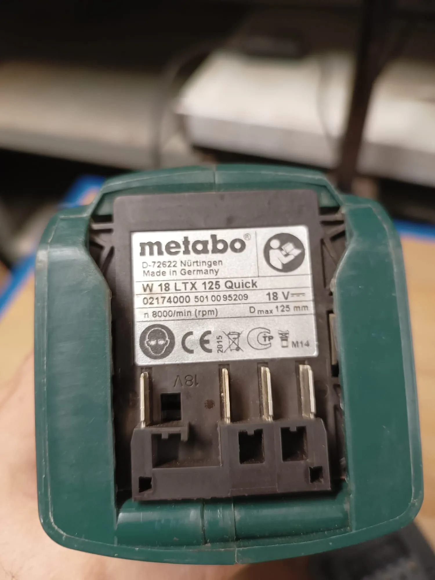 Zbliżenie na tabliczkę znamionową zielonego urządzenia marki Metabo. Na białym tle widnieje logo 'metabo', tekst 'D-72622 Nürtingen Made in Germany', model 'W 18 LTX 125 Quick', numer seryjny '02174000 5010095209', parametry 'n 8000/min (rpm)', '18 V' oraz 'Dmax 125 mm'. Poniżej znajdują się symbole certyfikacji, w tym znak CE oraz oznaczenie M14. Pod tabliczką widoczne są metalowe styki elektryczne w czarnej obudowie.