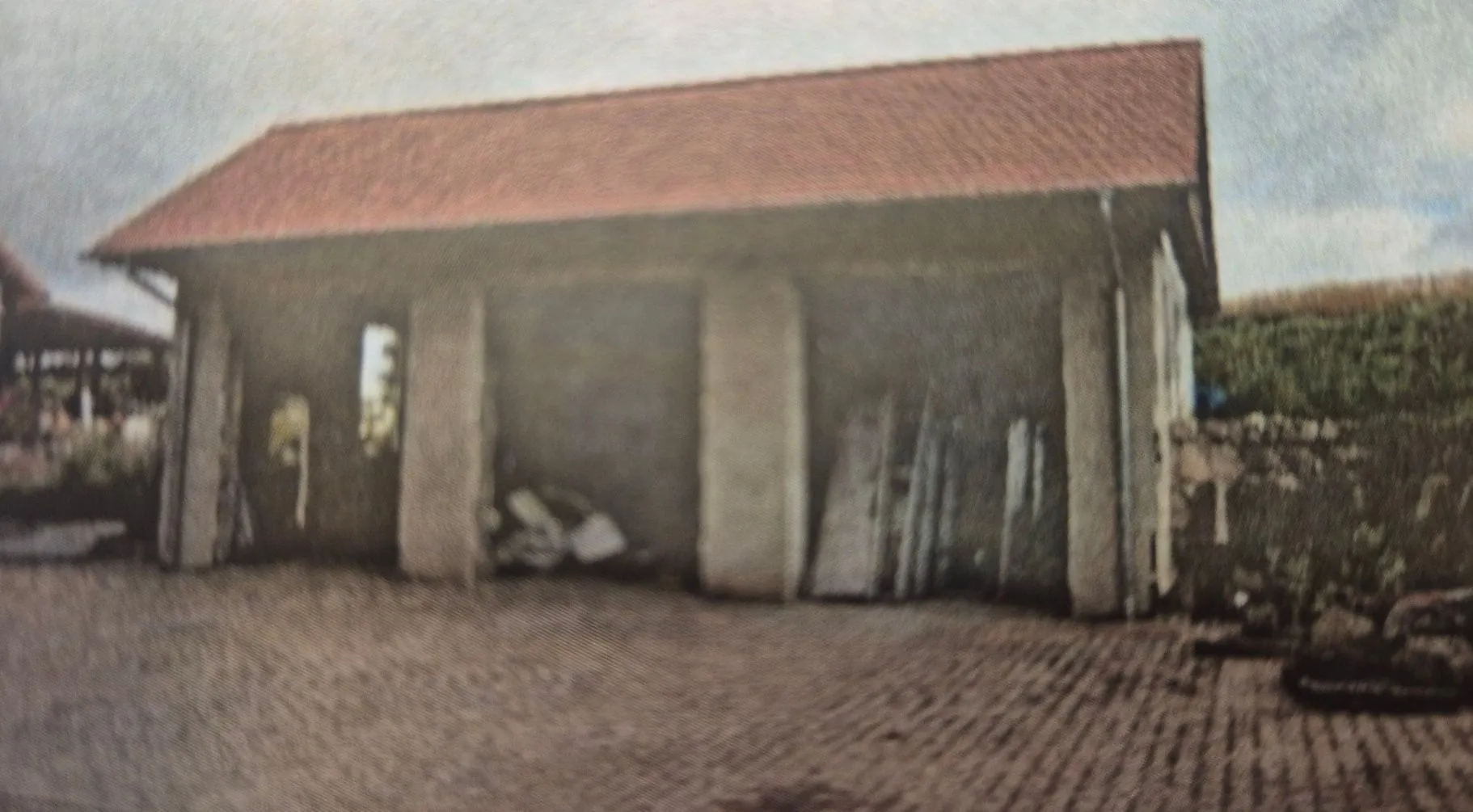 Zdjęcie przedstawiające prosty, betonowy budynek gospodarczy lub garaż z czerwonym, spadzistym dachem. Budynek ma otwarte wnęki wsparte na betonowych filarach, w których widoczne są różne przedmioty i materiały. Przed budynkiem znajduje się utwardzony plac z kostki brukowej.
