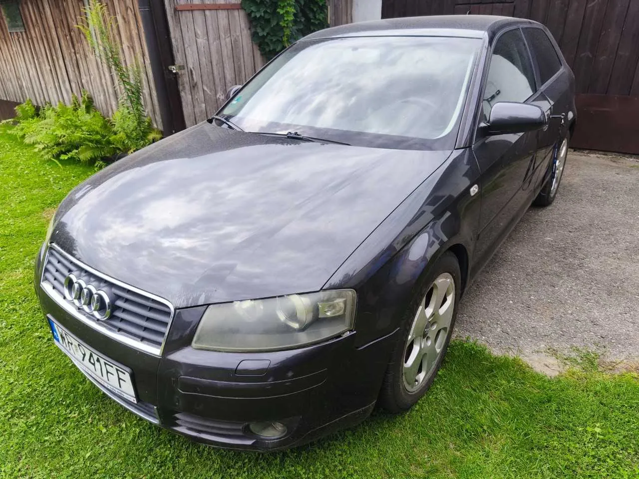 Ciemnofioletowy samochód marki Audi zaparkowany na podjeździe z betonu i trawy. Widoczny jest przód pojazdu z charakterystycznym grillem z czterema pierścieniami oraz tablicą rejestracyjną o numerze WR 941FF. W tle znajduje się drewniany płot i zielone rośliny.