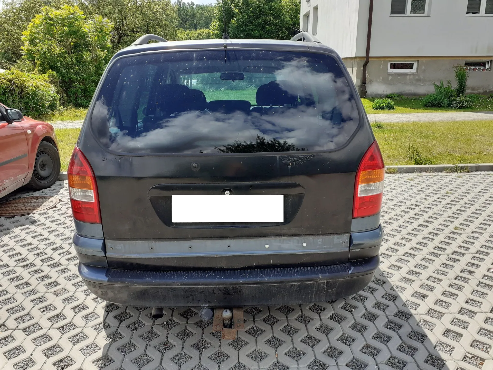 Tylny widok czarnego samochodu typu kombi zaparkowanego na kostce brukowej. Pojazd posiada zamontowany hak holowniczy. W tle widoczny jest fragment białego budynku oraz zielone drzewa i krzewy. Po lewej stronie zdjęcia widoczny jest fragment innego, czerwonego samochodu.