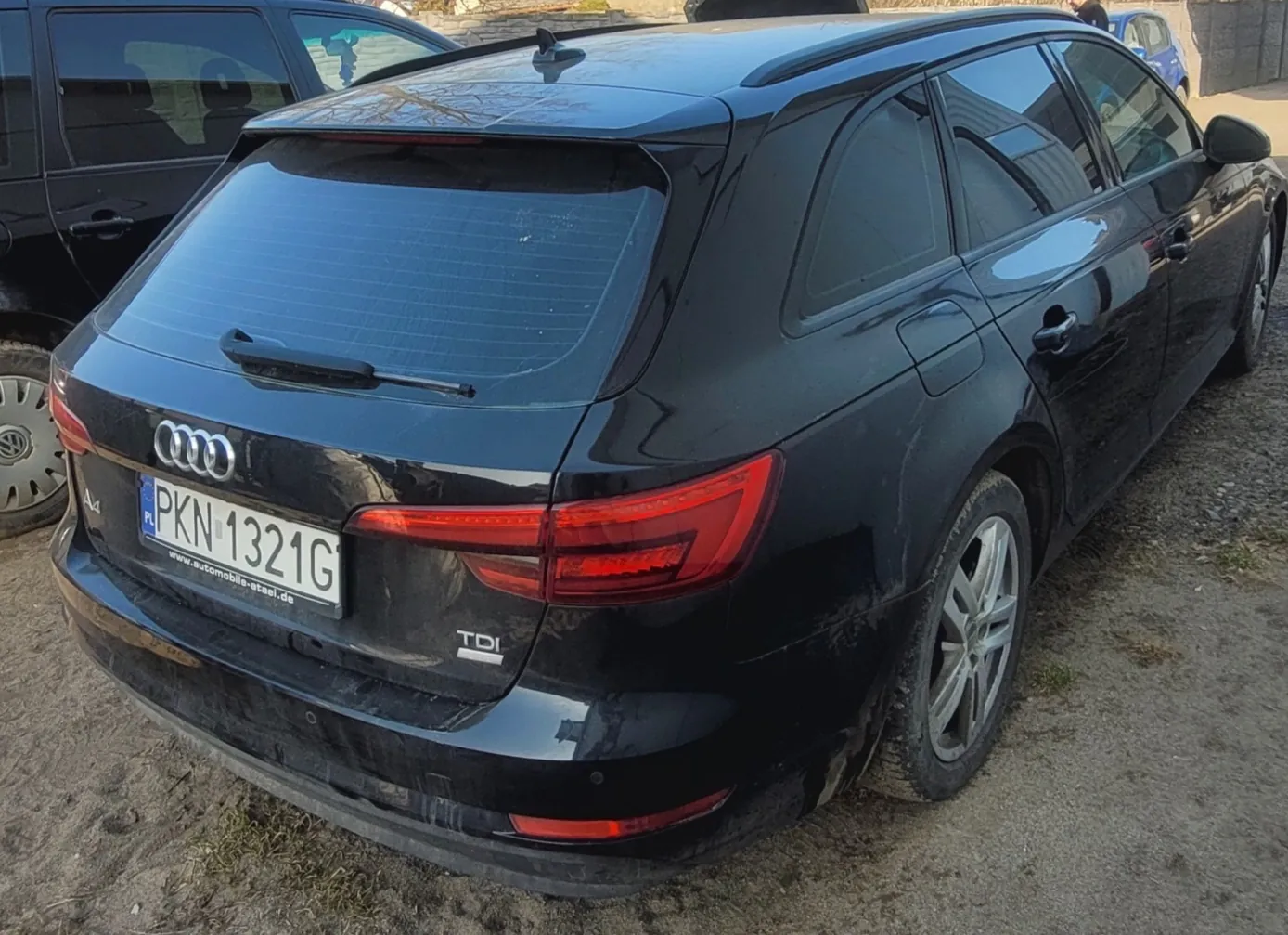 Czarny samochód Audi A4 Avant z widocznym tyłem. Na tablicy rejestracyjnej widnieje numer PKN 1321G oraz kod kraju PL. Poniżej tablicy znajduje się adres www.automobile-atael.de. Na bagażniku widoczne są logo Audi, oznaczenie A4 oraz TDI. Samochód stoi na podłożu z żwiru, w tle częściowo widoczne są inne pojazdy. Oświetlenie wskazuje na dzień.