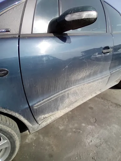 Boczny widok ciemnoniebieskiego samochodu z widocznymi zadrapaniami na drzwiach. Lusterko boczne z kierunkowskazem, brudna dolna część nadwozia. Na przednim błotniku widoczny napis 'ASSIC'. Samochód zaparkowany na betonowej powierzchni.