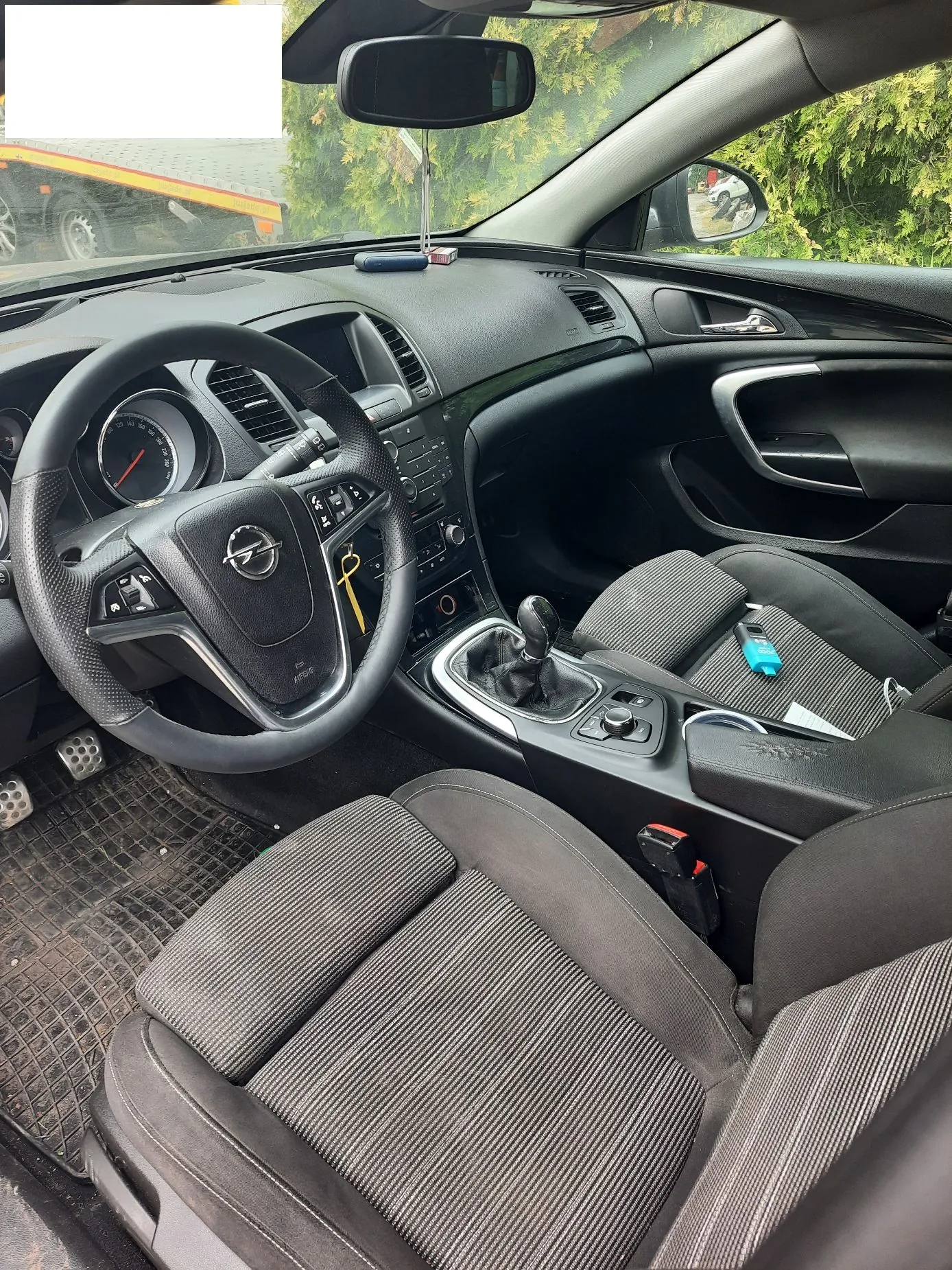 Wnętrze samochodu marki Opel widziane z perspektywy tylnego siedzenia. Widoczna jest czarna kierownica z logo producenta, deska rozdzielcza, dźwignia zmiany biegów oraz dwa przednie fotele z ciemnoszarą, prążkowaną tapicerką. Na siedzeniu pasażera leży niebieski przedmiot. Przez przednią szybę i lusterka widoczny jest fragment otoczenia z drzewami i pojazdem.