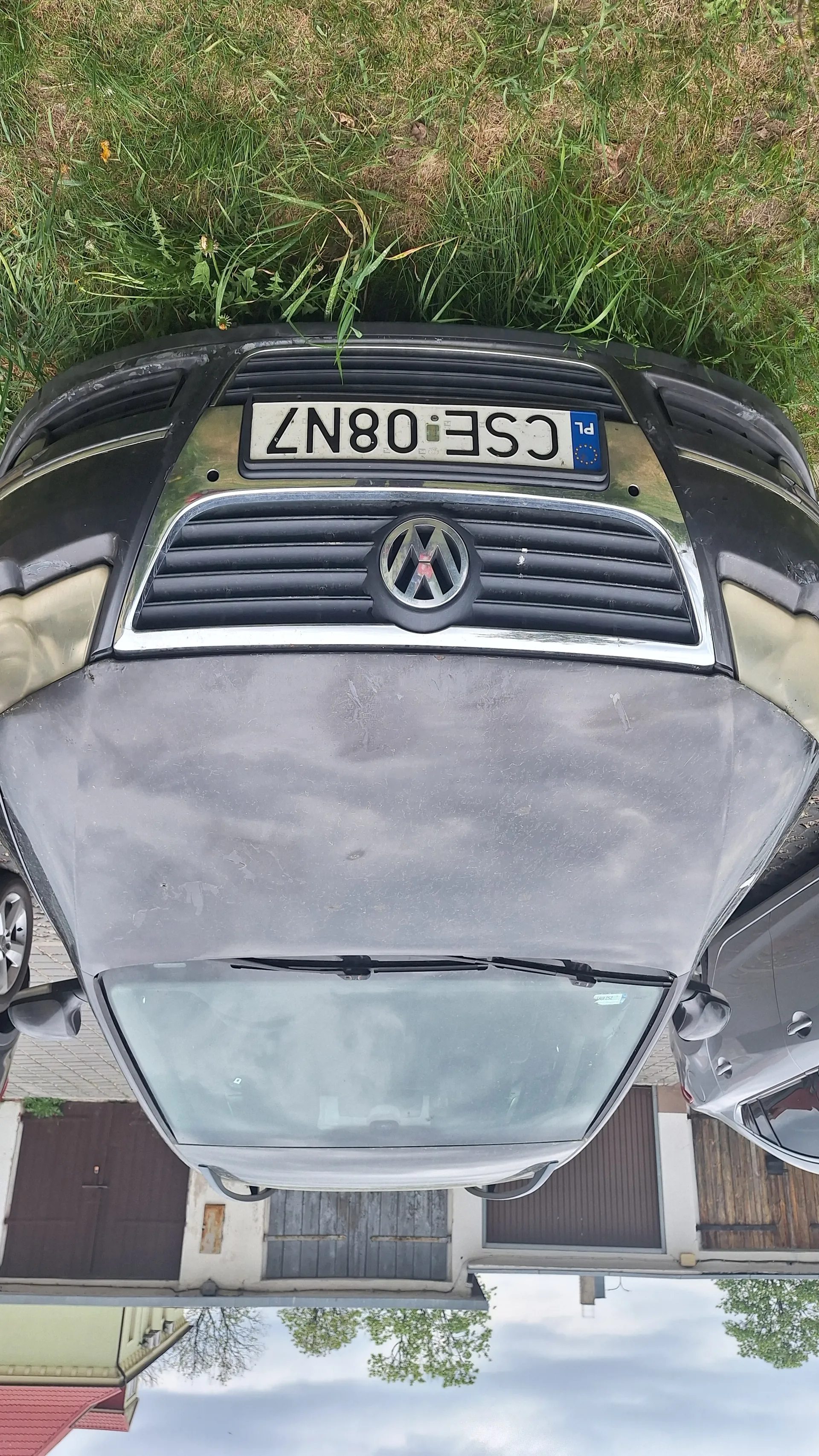 Czarny samochód marki Volkswagen z tablicą rejestracyjną CSE 08N7, widok z przodu. Samochód jest zabrudzony, widoczne logo VW na masce. Po prawej stronie trawa, w tle budynek z brązowymi drzwiami i czerwonym dachem. Niebo jest zachmurzone. Częściowo widoczny inny samochód.