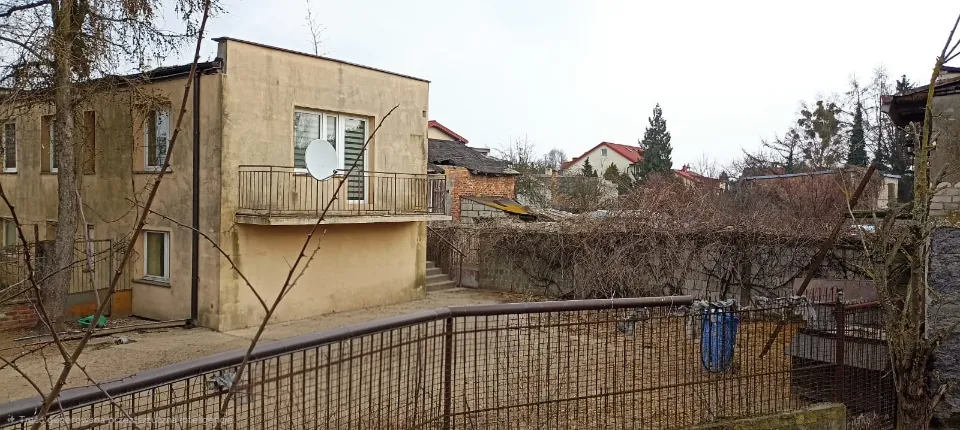 Zdjęcie przedstawiające zaplecze starego, beżowego budynku mieszkalnego z balkonem i białą anteną satelitarną. Obok budynku znajduje się ceglany budynek gospodarczy oraz betonowy mur porośnięty suchymi krzewami. Na pierwszym planie widoczny jest metalowy płot z siatką. W tle widać dachy innych domów i bezlistne drzewa pod zachmurzonym niebem.