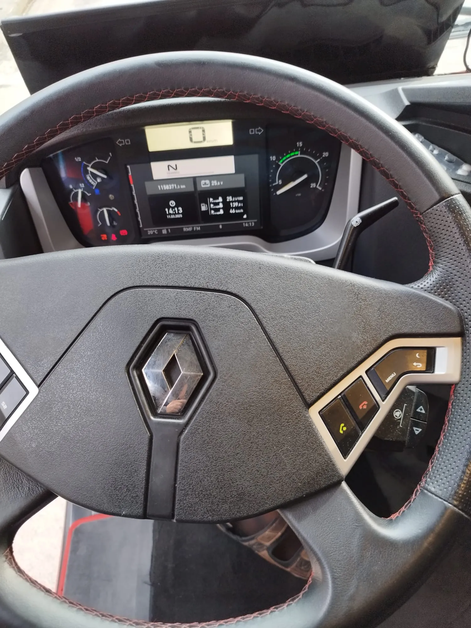 Kierownica samochodu marki Renault z czarnym obiciem i czerwonym przeszyciem. W centrum kierownicy znajduje się logo Renault w kształcie rombu. Na kierownicy widoczne są przyciski sterujące, w tym żółty przycisk telefonu i przyciski nawigacyjne. Za kierownicą umieszczony jest cyfrowy zegar pokazujący prędkość 0 km/h, bieżący przebieg 1158371,3 km, godzinę 14:13, datę 11.03.2023 oraz temperaturę 20°C. Wskazówka biegu wskazuje pozycję N (neutral). Po prawej stronie zegara znajduje się tachometr z zielonym segmentem. W tle widoczna jest czarna deska rozdzielcza.
