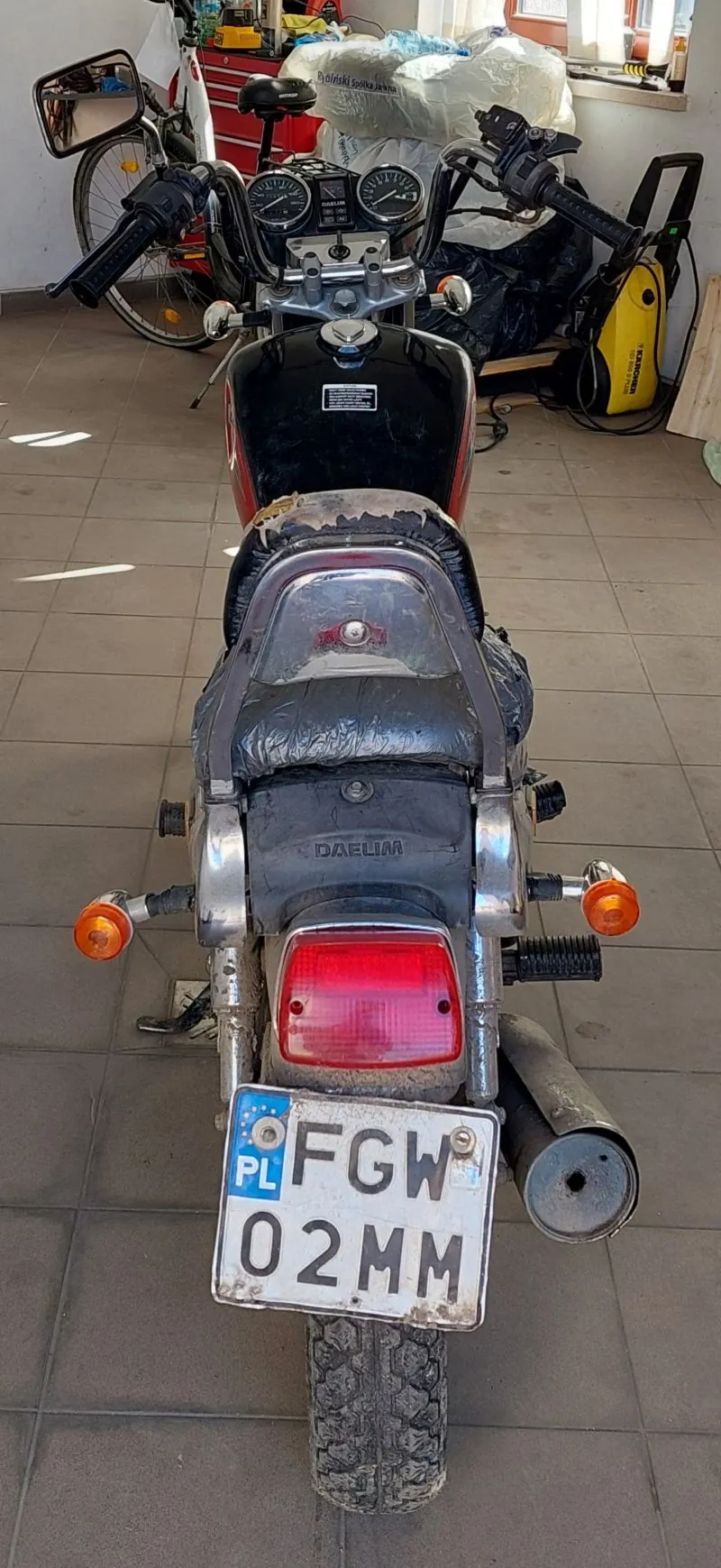Zdjęcie przedstawia motocykl marki Daelim widziany od tyłu. Na motocyklu widoczna jest tablica rejestracyjna z napisem PL FGW 02MM, czerwone światło tylnego reflektora oraz pomarańczowe kierunkowskazy. Siedzenie jest czarne, częściowo zniszczone, a na baku znajduje się czarna tabliczka z białym napisem. W tle widoczne są elementy garażu: rower, czerwony kufer narzędziowy, żółty odkurzacz oraz opakowania na półce. Podłoga wyłożona jest szarymi płytkami.