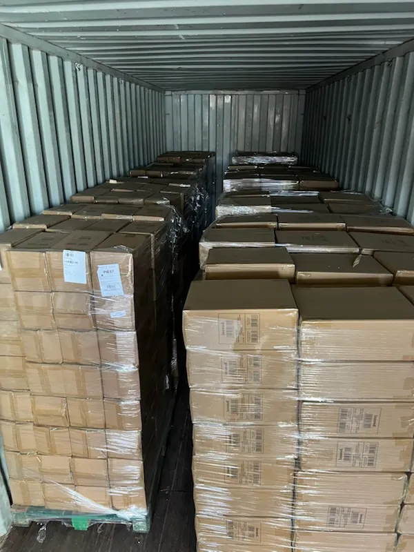 Wnętrze kontenera transportowego wypełnione stosami brązowych kartonów owiniętych przezroczystą folią. Kartony są ułożone w rzędach, niektóre mają białe etykiety z nieczytelnym tekstem. Ściany kontenera są metalowe, pionowe, z widocznymi listwami. Podłoga kontenera jest ciemna, prawdopodobnie drewniana lub metalowa.
