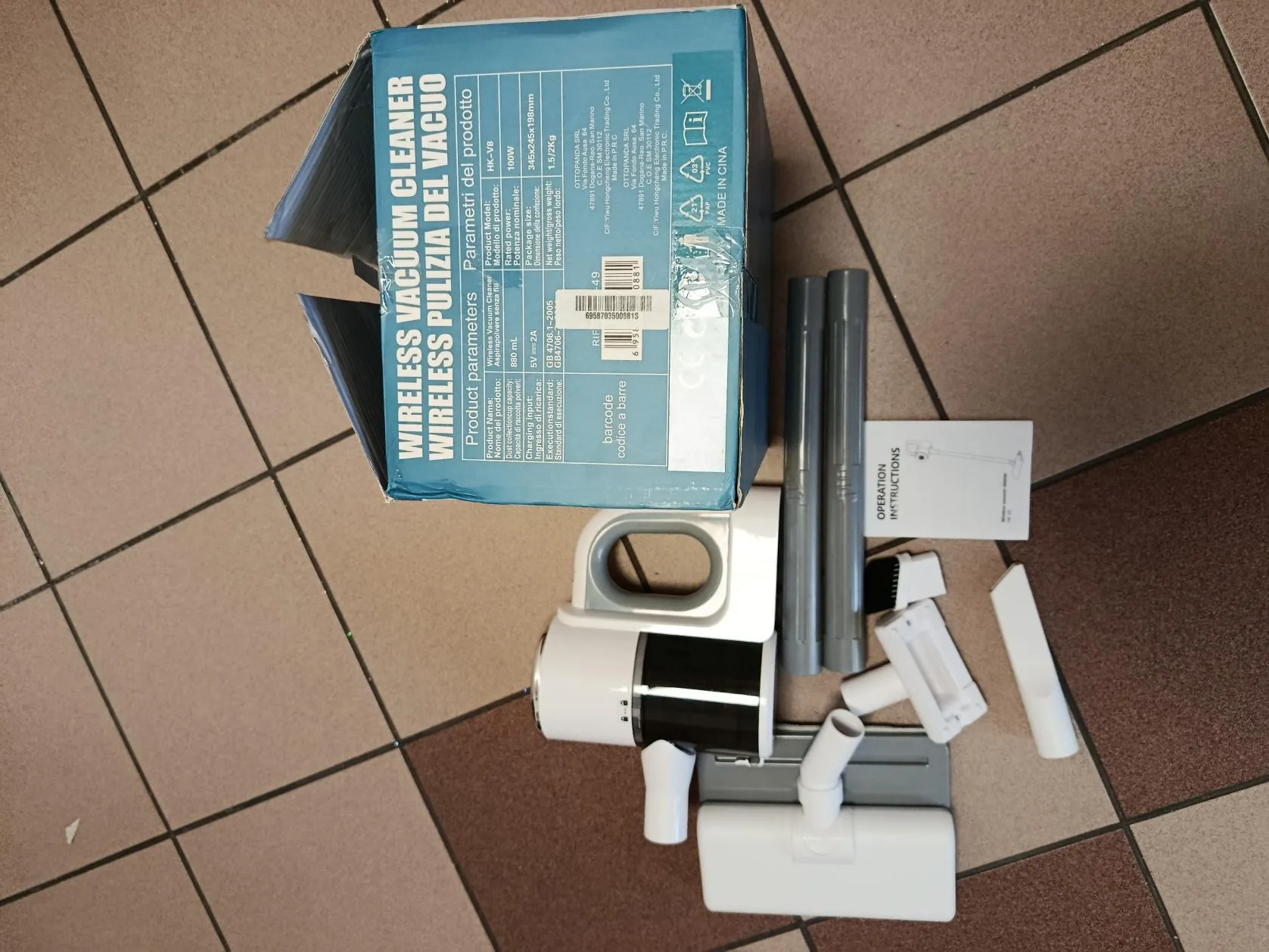 Zdjęcie przedstawia zestaw bezprzewodowego odkurzacza na podłodze z kafelkami w brązowo-beżowych odcieniach. W centrum znajduje się niebieskie pudełko z napisem 'WIRELESS VACUUM CLEANER' oraz 'PULIZIA DEL VACUO' w białych literach. Obok pudełka leży biało-szary odkurzacz ręczny z czarnym akumulatorem, dwa szare rury, białe akcesoria (w tym szczotka i końcówki), oraz instrukcja z napisem 'OPERATION INSTRUCTIONS'. Na pudełku widoczne są parametry techniczne, kod kreskowy i informacja 'MADE IN CHINA'.