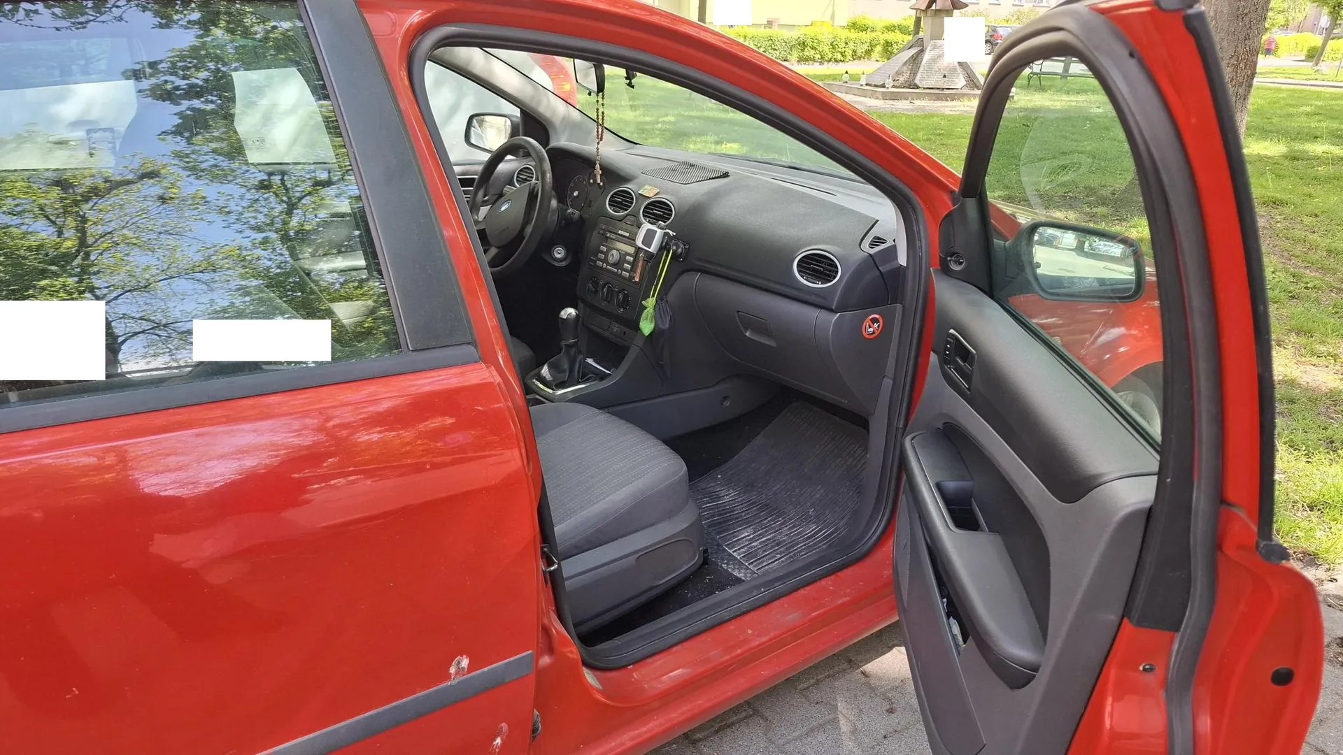 Zbliżenie na otwarte drzwi kierowcy w czerwonym samochodzie. Widoczny jest fragment nadwozia, wnętrze pojazdu z czarną tapicerką, kierownicą, deską rozdzielczą oraz otwarta drzwi z lusterkiem bocznym. W tle widać zieloną trawę i drzewa.