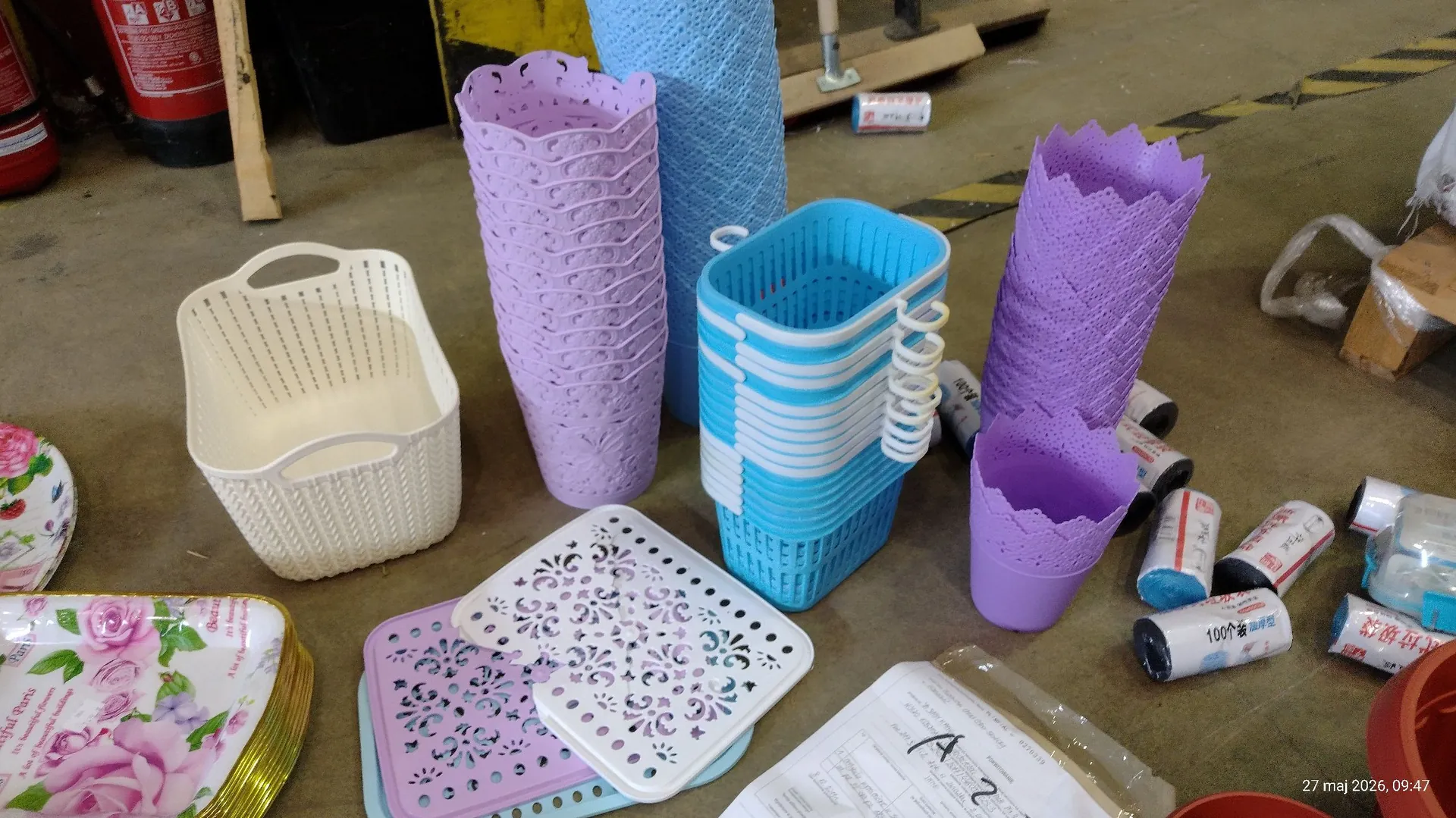 Kolekcja plastikowych przedmiotów codziennego użytku rozrzuconych na szarej, betonowej podłodze. Widoczne są: biały ażurowy koszyk, dwa wysokie fioletowe koszyki o dekoracyjnym splocie, niebieski ażurowy koszyk oraz niebieski walec z plastiku. Obok znajdują się dwie białe, ażurowe tace z wycinanymi wzorami, jedna z nich leży na fioletowej tacy. Po lewej stronie widoczny jest fragment tacy z nadrukiem różowych kwiatów. Po prawej stronie leży kilka białych i niebieskich cylindrycznych opakowań. W tle widoczne są fragmenty drewnianych desek i czerwone gaśnice.