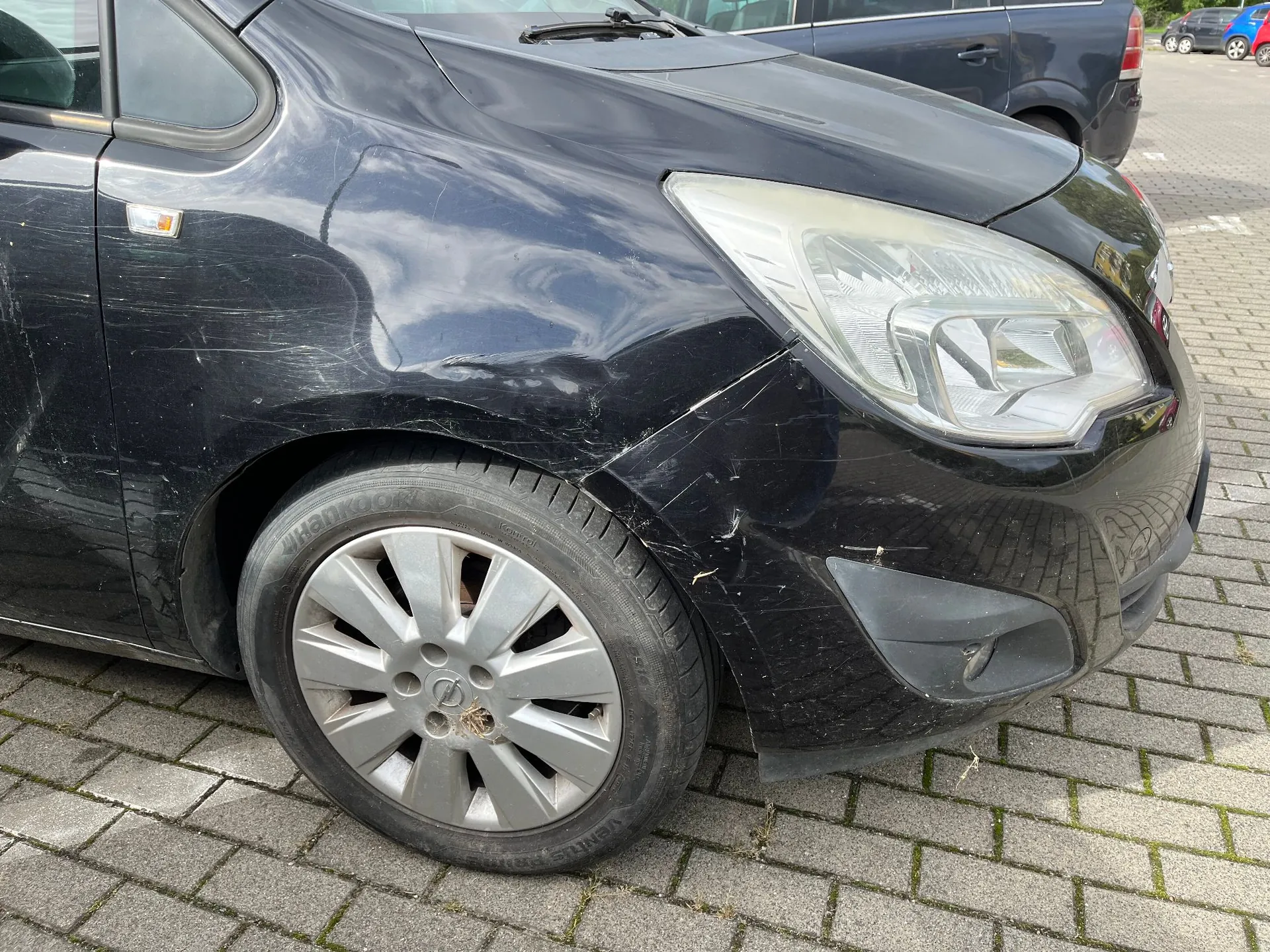 Zbliżenie na przedni prawy narożnik czarnego samochodu zaparkowanego na kostce brukowej. Widoczne są uszkodzenia karoserii w postaci wgniecenia i zarysowań na błotniku oraz zderzaku, tuż przed reflektorem. Widoczna jest srebrna felga z kołpakiem i opona marki Hankook.
