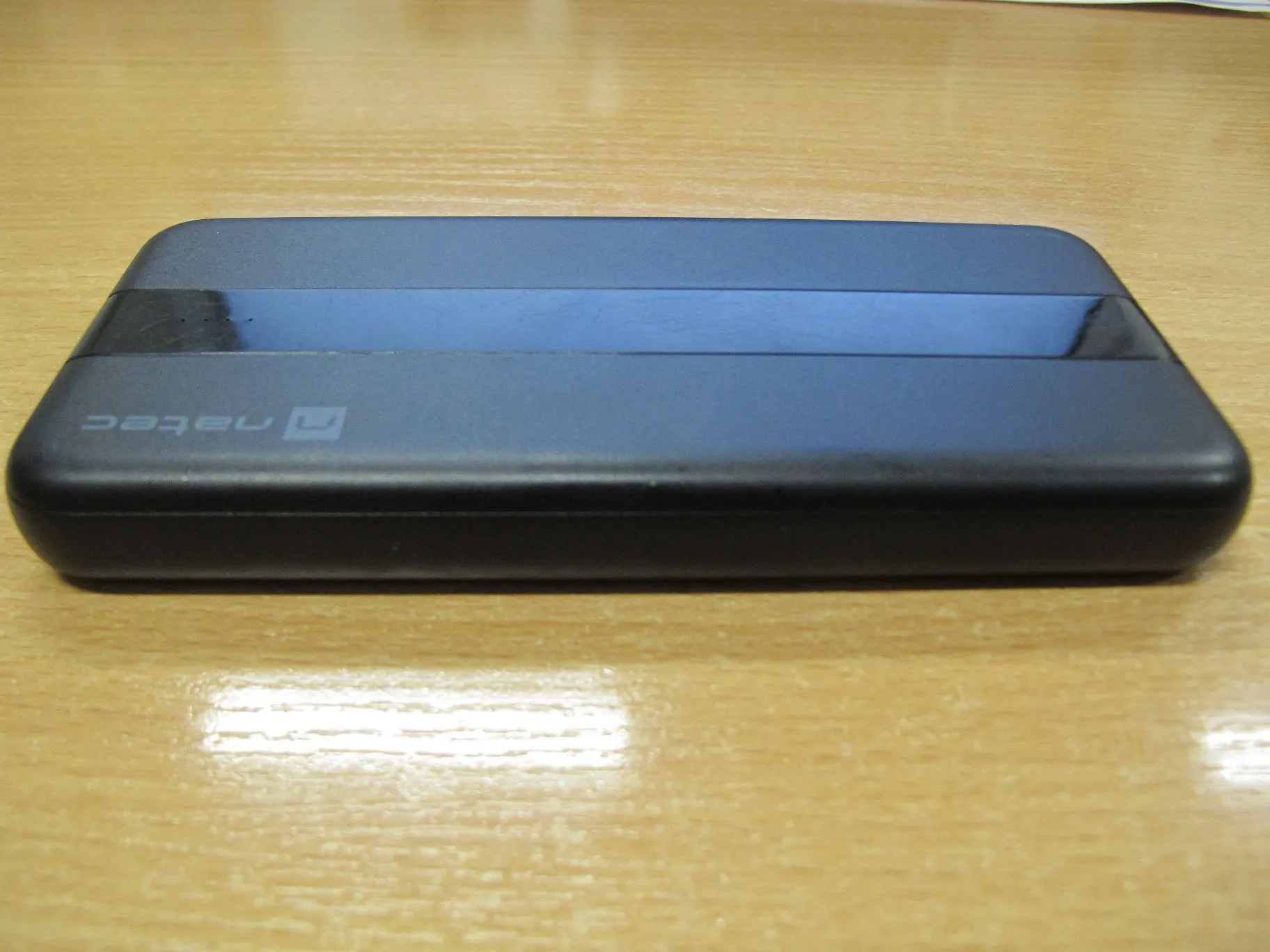 Czarny, prostokątny powerbank marki natec leżący poziomo na jasnym, drewnianym blacie. Na obudowie urządzenia widoczne jest logo i napis 'natec'.