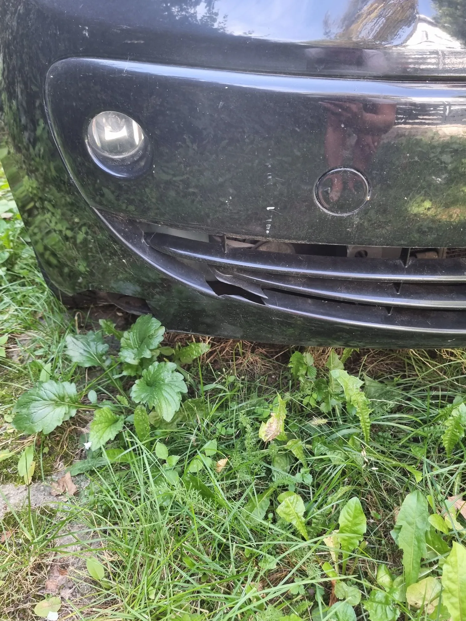 Zbliżenie na przedni, czarny zderzak samochodu, który jest uszkodzony. W dolnej części zderzaka widoczne jest wgniecenie i pęknięcie plastiku. Po lewej stronie znajduje się okrągła lampka przeciwmgielna, a obok niej mały, okrągły otwór serwisowy. Samochód stoi na zielonej trawie z chwastami.