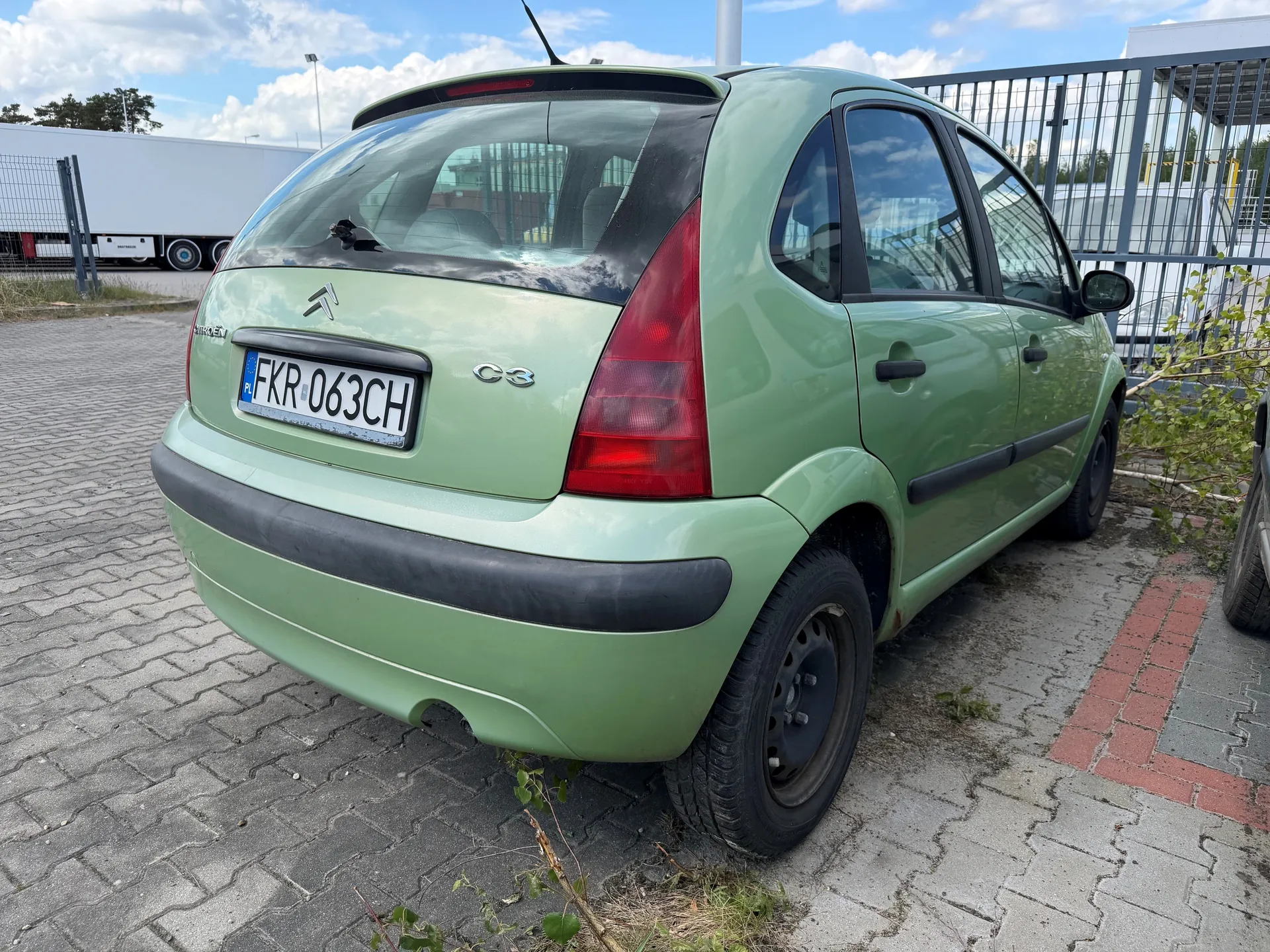 Zielony samochód Citroën C3 zaparkowany na brukowanej powierzchni. Widoczny tył pojazdu z tablicą rejestracyjną FKR-063CH oraz oznaczeniem PL. W tle biały naczepa ciężarowa, metalowe ogrodzenie, budynki i niebo z chmurami. Na ziemi rosną pojedyncze rośliny. Samochód ma czarne felgi i czerwone światła tylnego. Oznaczenie C3 znajduje się po prawej stronie bagażnika.