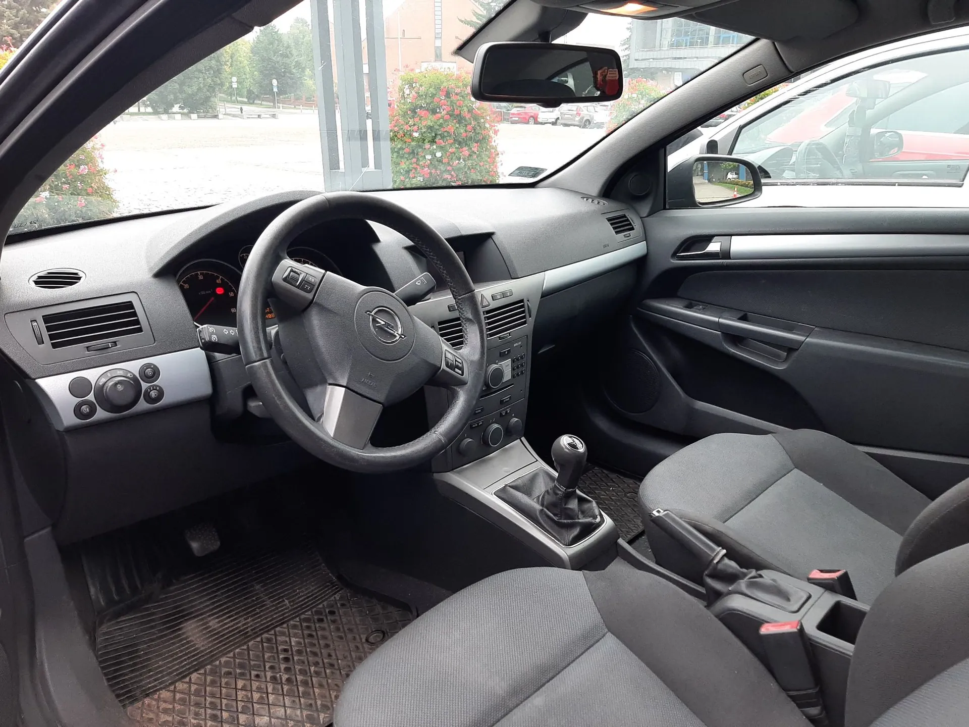 Wnętrze samochodu widziane z perspektywy kierowcy. Widoczna jest czarna kierownica z logo marki Opel, deska rozdzielcza z zegarami, system audio oraz lewarek zmiany biegów. Fotele są obite szarym materiałem. Przez przednią szybę i lusterka widać fragmenty otoczenia zewnętrznego.