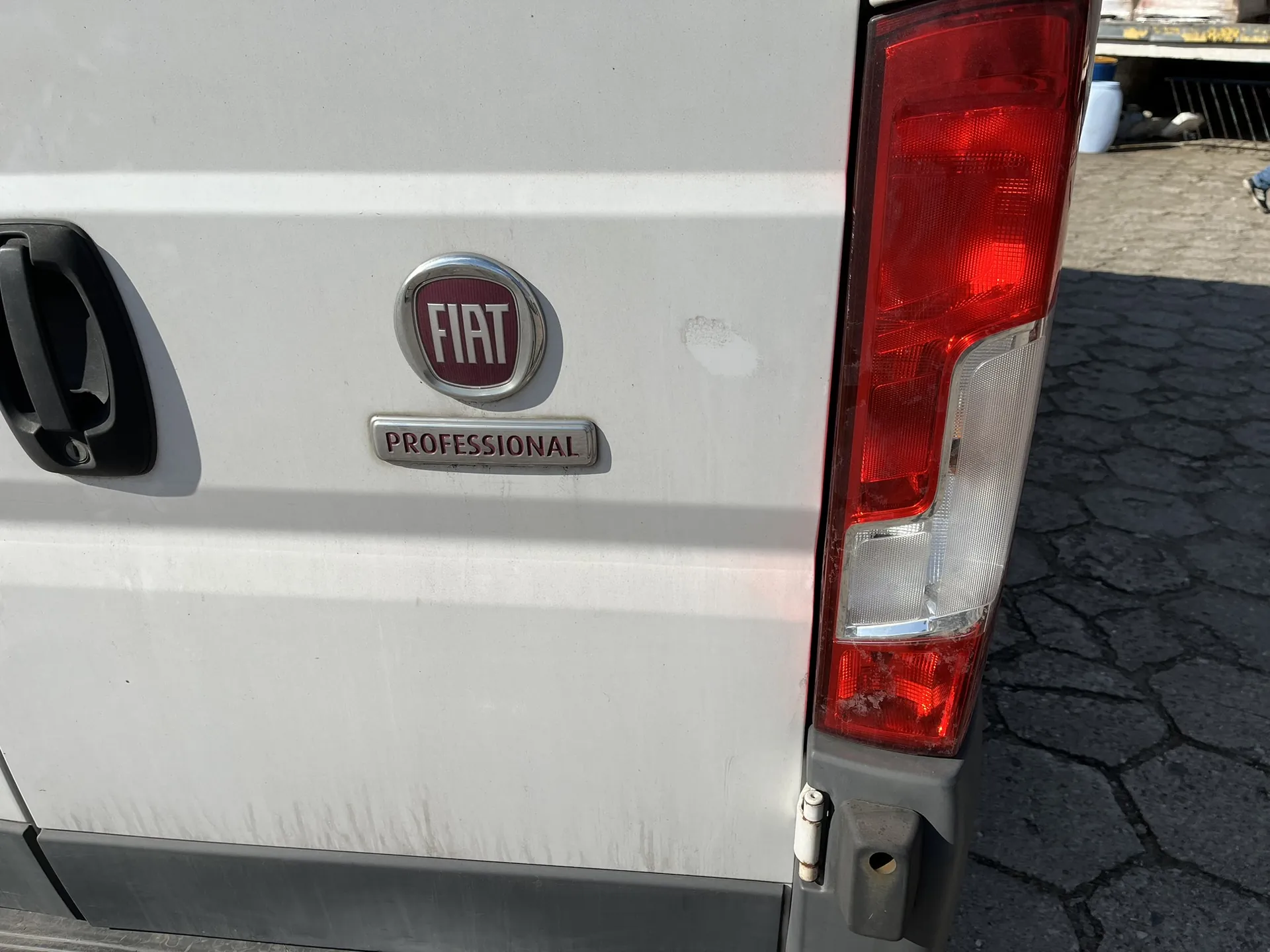 Zbliżenie na tylną część białego samochodu marki Fiat. Widoczne jest czerwone, okrągłe logo FIAT oraz poniżej srebrna tabliczka z napisem PROFESSIONAL. Po prawej stronie znajduje się pionowa lampa tylna w kolorze czerwonym i białym. W lewej części obrazu widoczny jest fragment czarnej klamki. Samochód stoi na szarej, kostce brukowej.