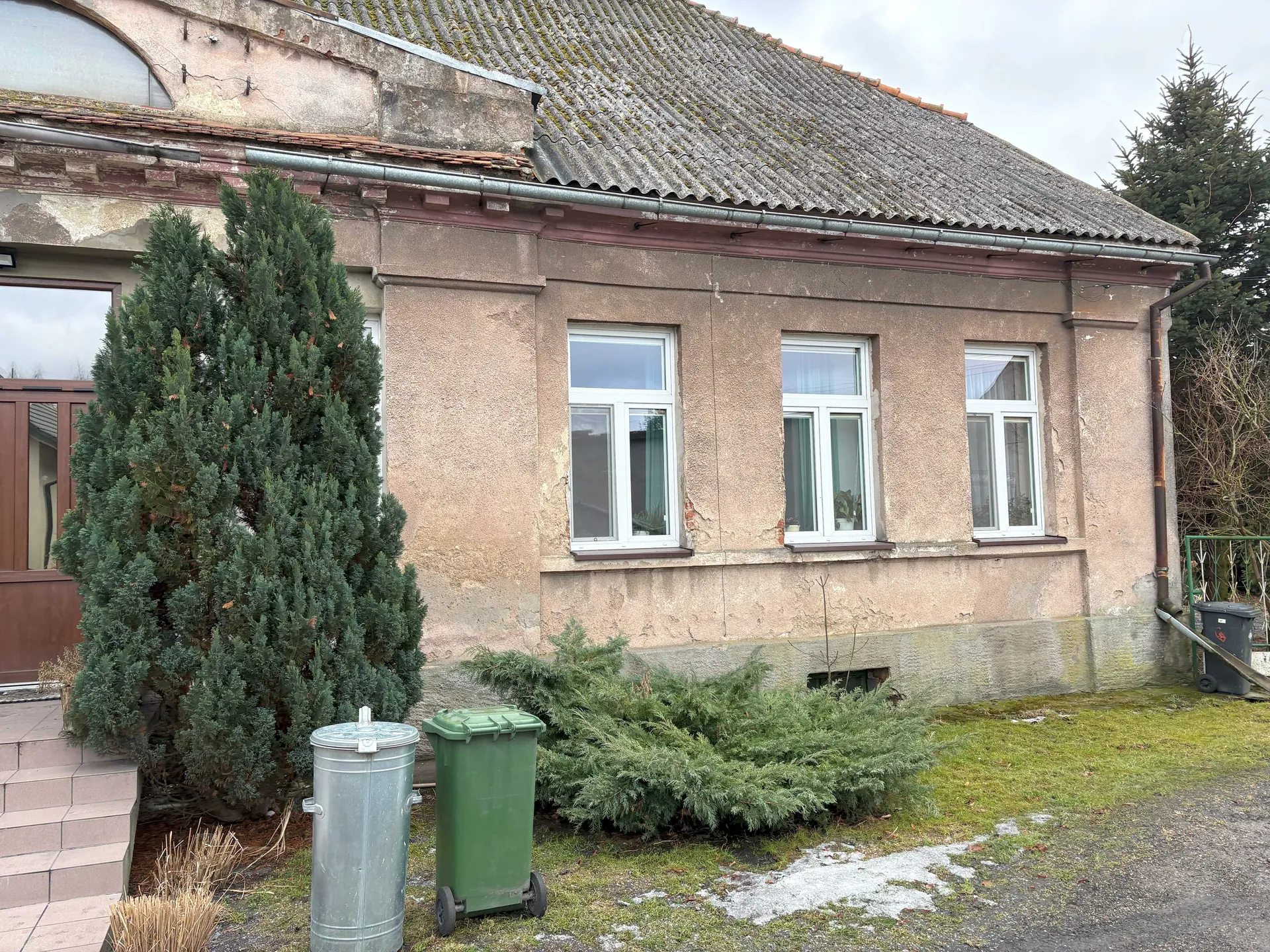 Zdjęcie przedstawia jednopiętrowy budynek z dachem krytym dachówką. Fasada budynku jest beżowa z widocznymi uszkodzeniami i odsłoniętymi fragmentami muru. Na ścianie znajdują się trzy białe okna z podwójnymi szybami. Po lewej stronie widoczne są schody prowadzące do brązowych drzwi oraz duża zielona krzewina. Przed budynkiem stoją dwa pojemniki na śmieci: srebrny metalowy i zielony na kółkach. Na trawniku leżą fragmenty śniegu lub lodu. W tle rosną drzewa iglaste i liściaste, a po prawej stronie widoczny jest czarny pojemnik na śmieci oraz zielone ogrodzenie. Niebo jest zachmurzone.