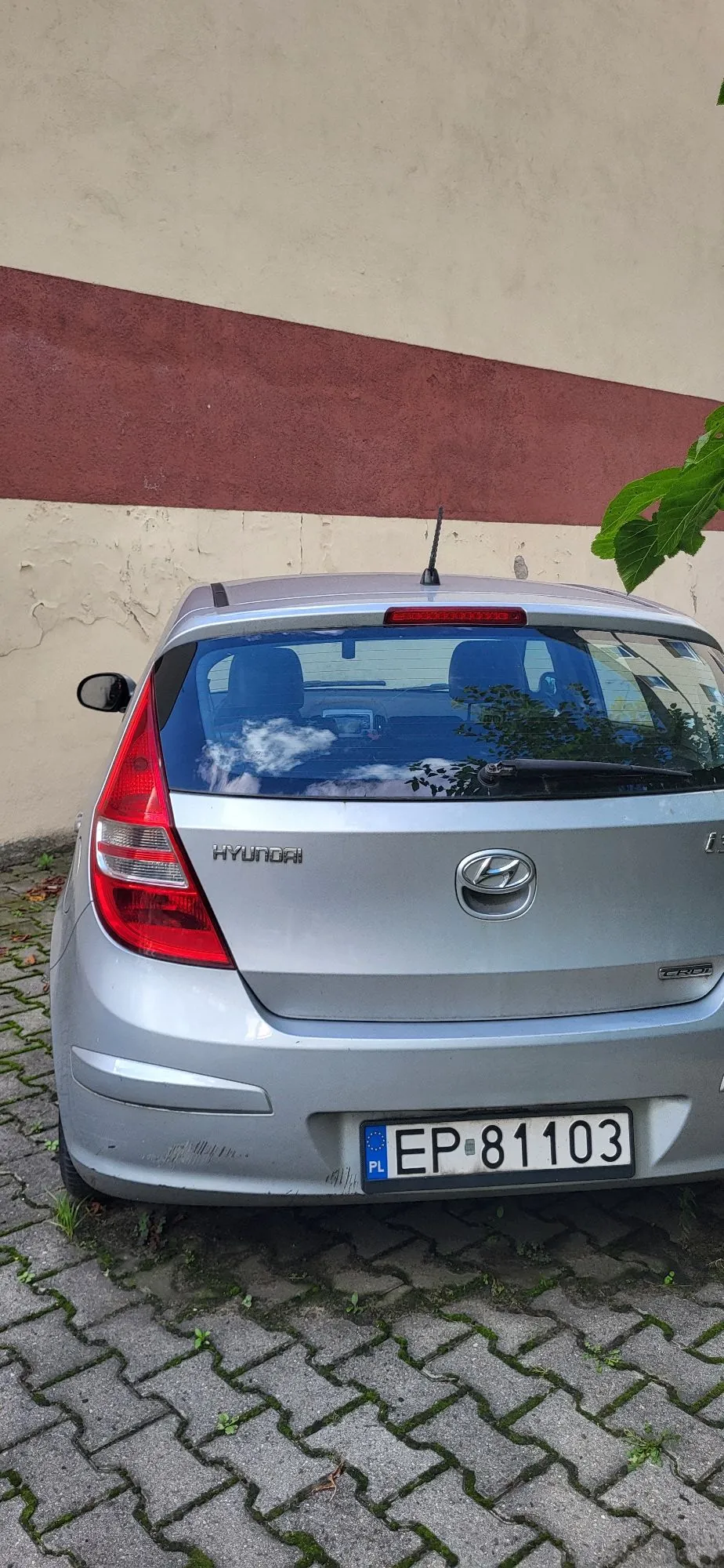 Srebrny samochód marki Hyundai zaparkowany tyłem do ściany budynku. Na klapie bagażnika widnieje napis HYUNDAI oraz logo marki. Samochód posiada polską tablicę rejestracyjną o numerze EP 81103. Podłoże stanowi szara kostka brukowa z mchem w szczelinach. Ściana w tle jest pomalowana na dwa kolory: beżowy i bordowy.