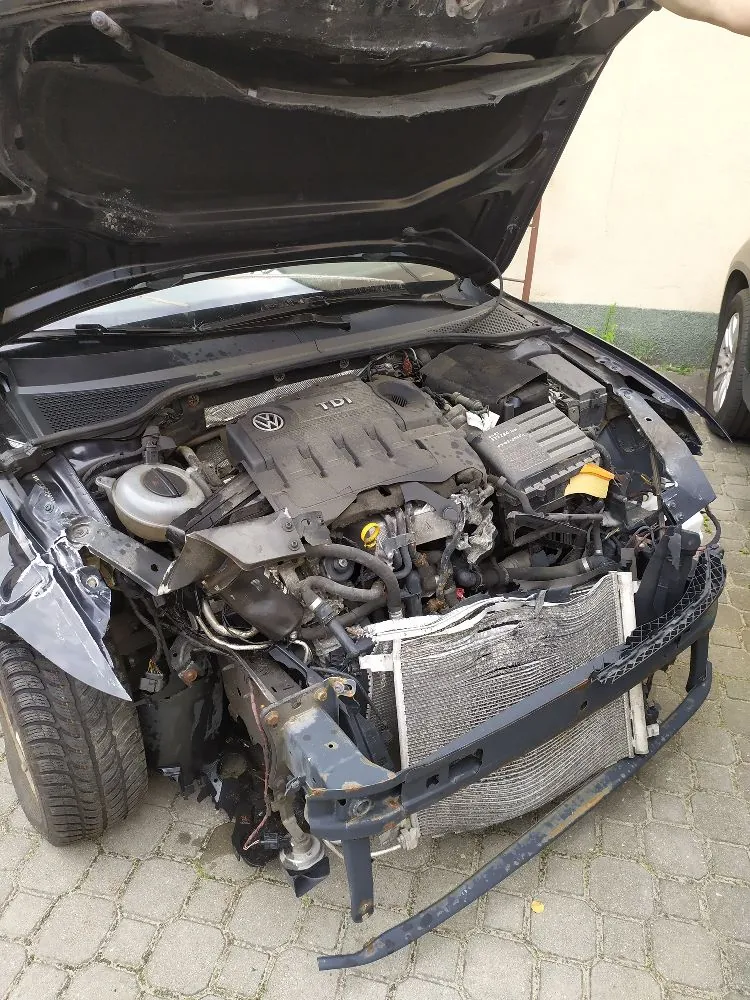 Zdjęcie przedstawiające uszkodzony przód ciemnego samochodu z otwartą maską, odsłaniającą silnik z logo Volkswagena i napisem TDI. Pojazd jest poważnie rozbity: brakuje przedniego zderzaka i grilla, widoczna jest zniekształcona rama oraz uszkodzony chłodnica. Lewe przednie skrzydło jest wgniecione.