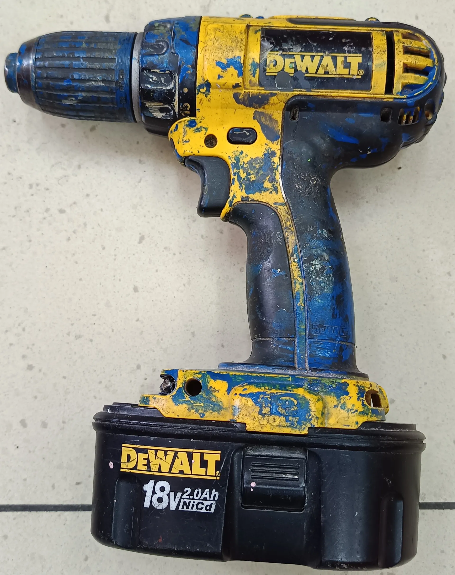 Zdjęcie przedstawia zużyty akumulatorowy wiertarko-wkrętarkę marki DeWALT. Urządzenie ma kolor żółto-niebieski z widocznymi śladami zużycia i obłupaną farbą. Na korpusie widnieje logo 'DeWALT', a na czarnym akumulatorze umieszczono napisy: 'DeWALT', '18V 2.0Ah NiCd'. Wiertarka leży na jasnej, kropkowanej powierzchni.