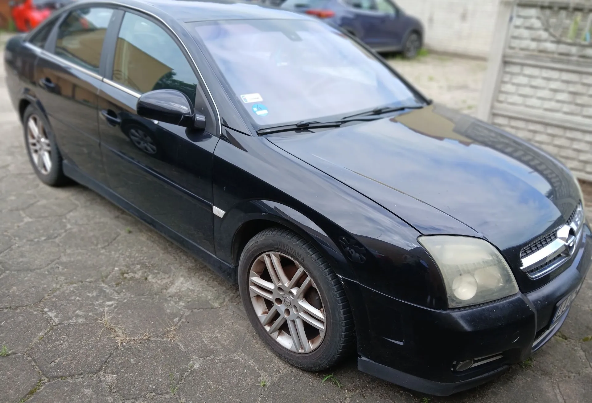 Czarny samochód osobowy typu sedan zaparkowany na kostce brukowej. Widok z przodu i z boku pojazdu. Samochód ma srebrne felgi aluminiowe i lekko zmatowione reflektory przednie.