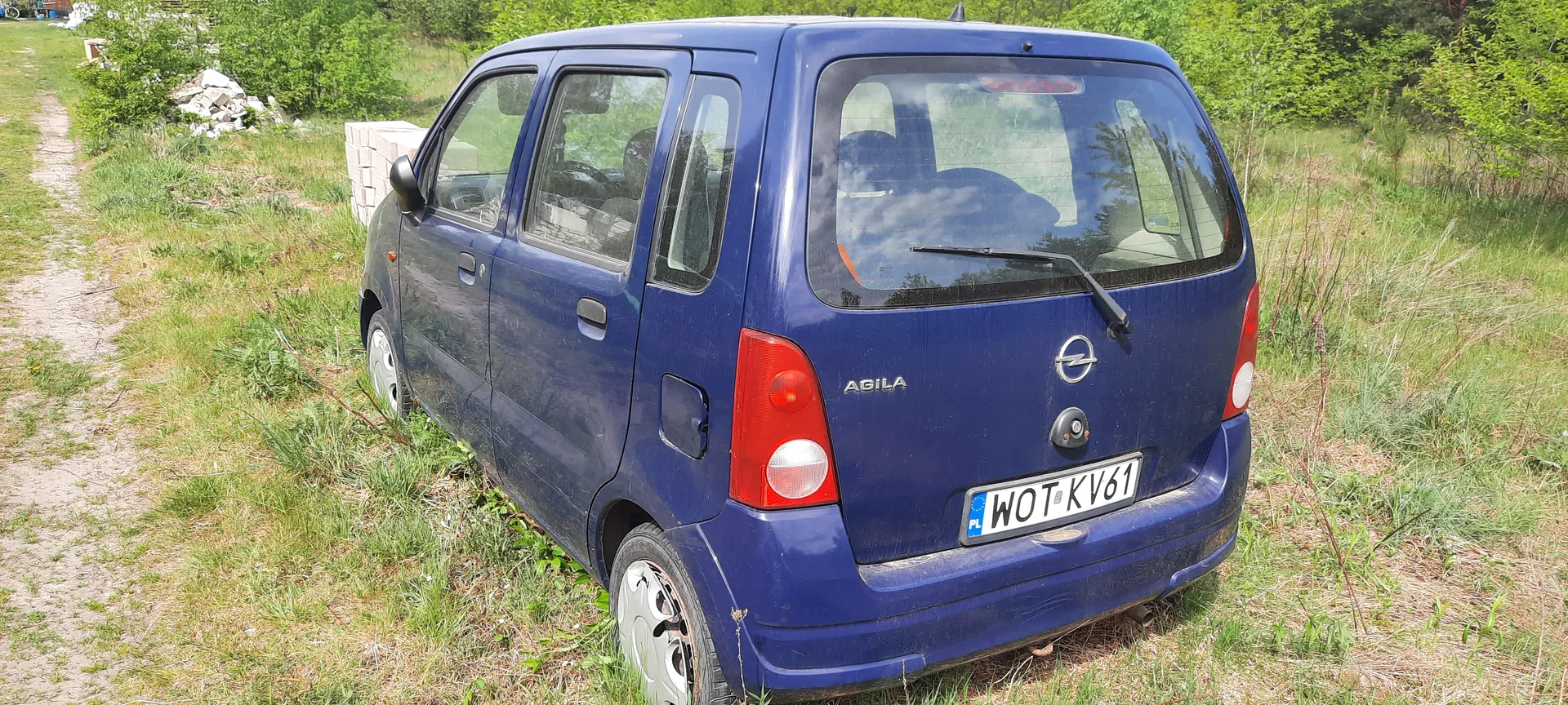 Niebieski samochód Opel Agila zaparkowany na trawie. Widoczny tył pojazdu z czerwonymi światłami, tablicą rejestracyjną PL WOT KV61 oraz napisem AGILA pod logo marki. W tle zielone krzewy, drzewa, stos białych cegieł i ziemna droga po lewej stronie. Samochód znajduje się na trawiastym terenie z suchą trawą i roślinnością.