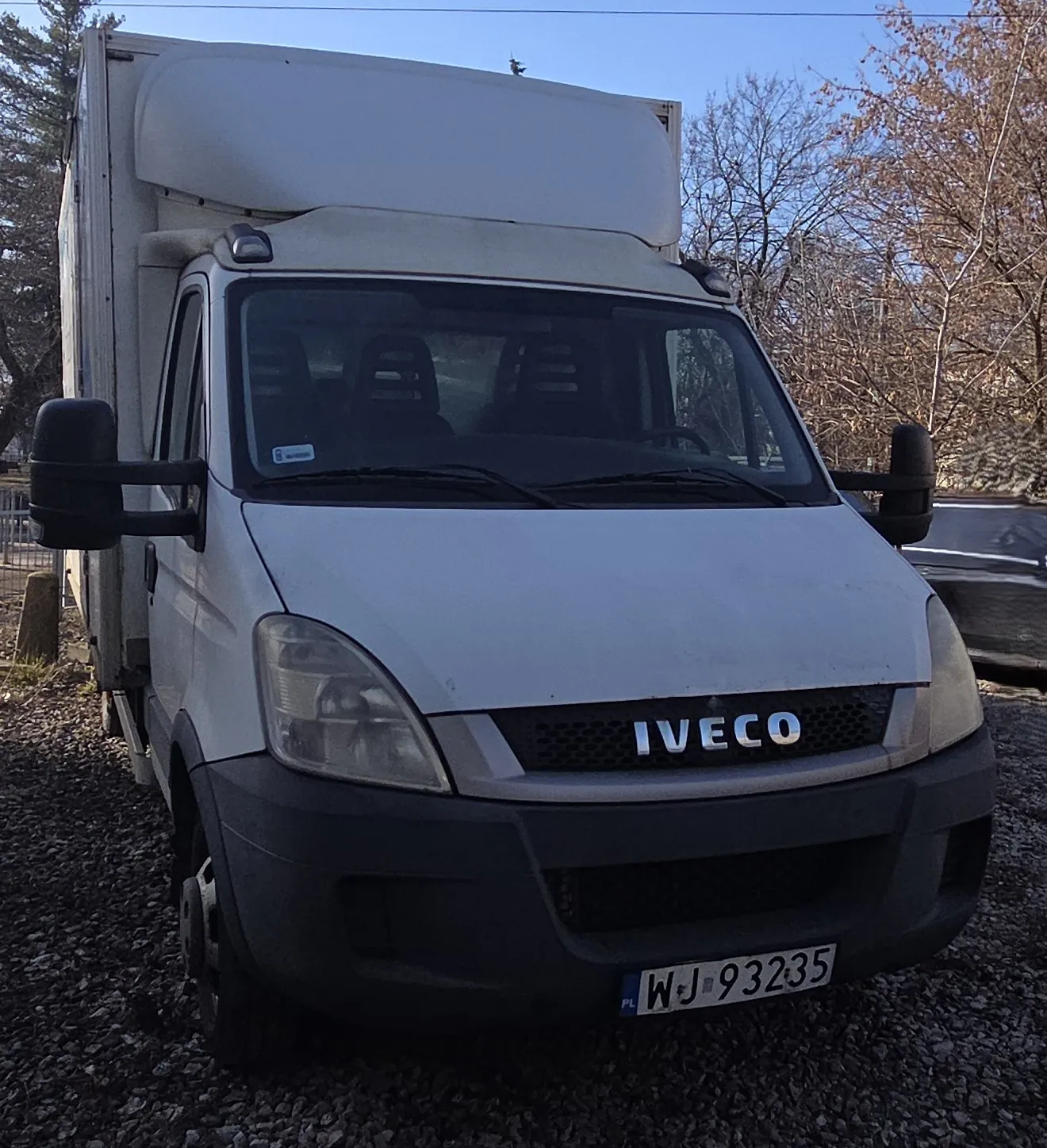 Biały samochód dostawczy marki IVECO z przyczepą, zaparkowany na żwirowym podłożu. Widoczna tablica rejestracyjna PL WJ 93235. Przednia szyba z czarnymi siedzeniami wewnątrz, lusterka boczne oraz logo IVECO na masce. Tło stanowią drzewa bez liści i jasne niebo. Samochód stoi prosto, z horyzontem równoległym do dolnej krawędzi zdjęcia.