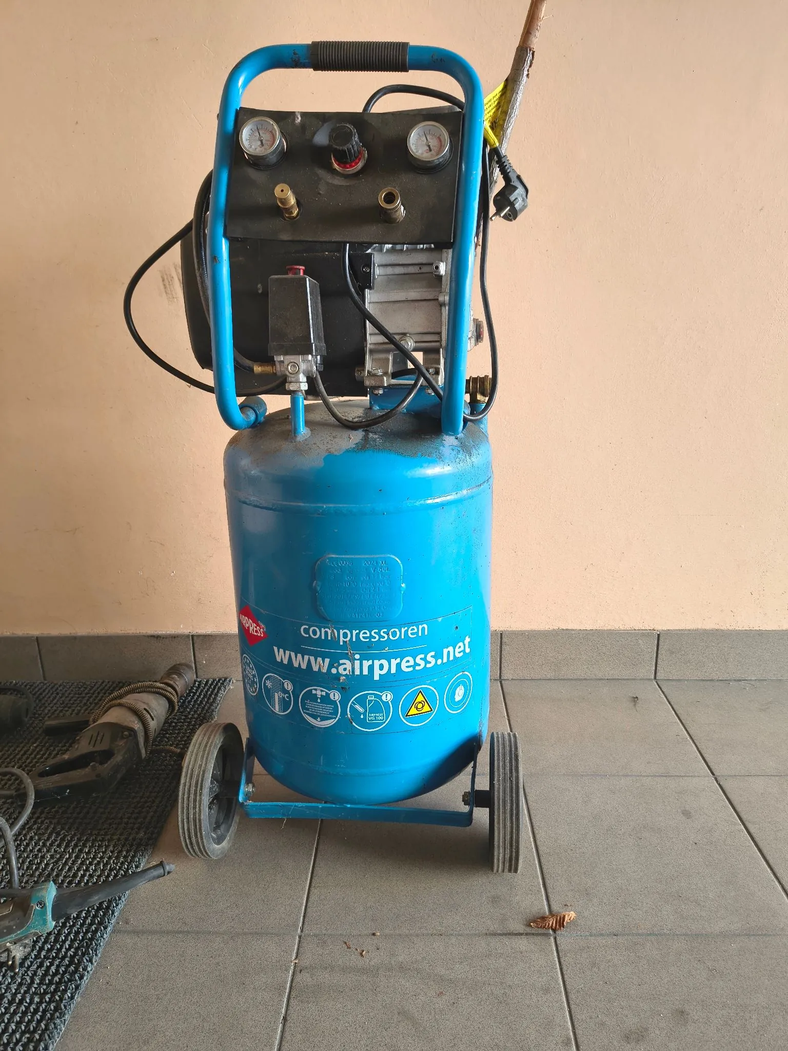 Niebieska sprężarka powietrza na kółkach, stojąca na szarych płytkach. Urządzenie posiada pionowy zbiornik z napisem 'compressoren www.airpress.net', górny panel z dwoma manometrami i zaworami oraz niebieską ramę z uchwytem. Obok sprężarki na ziemi leżą narzędzia elektryczne.