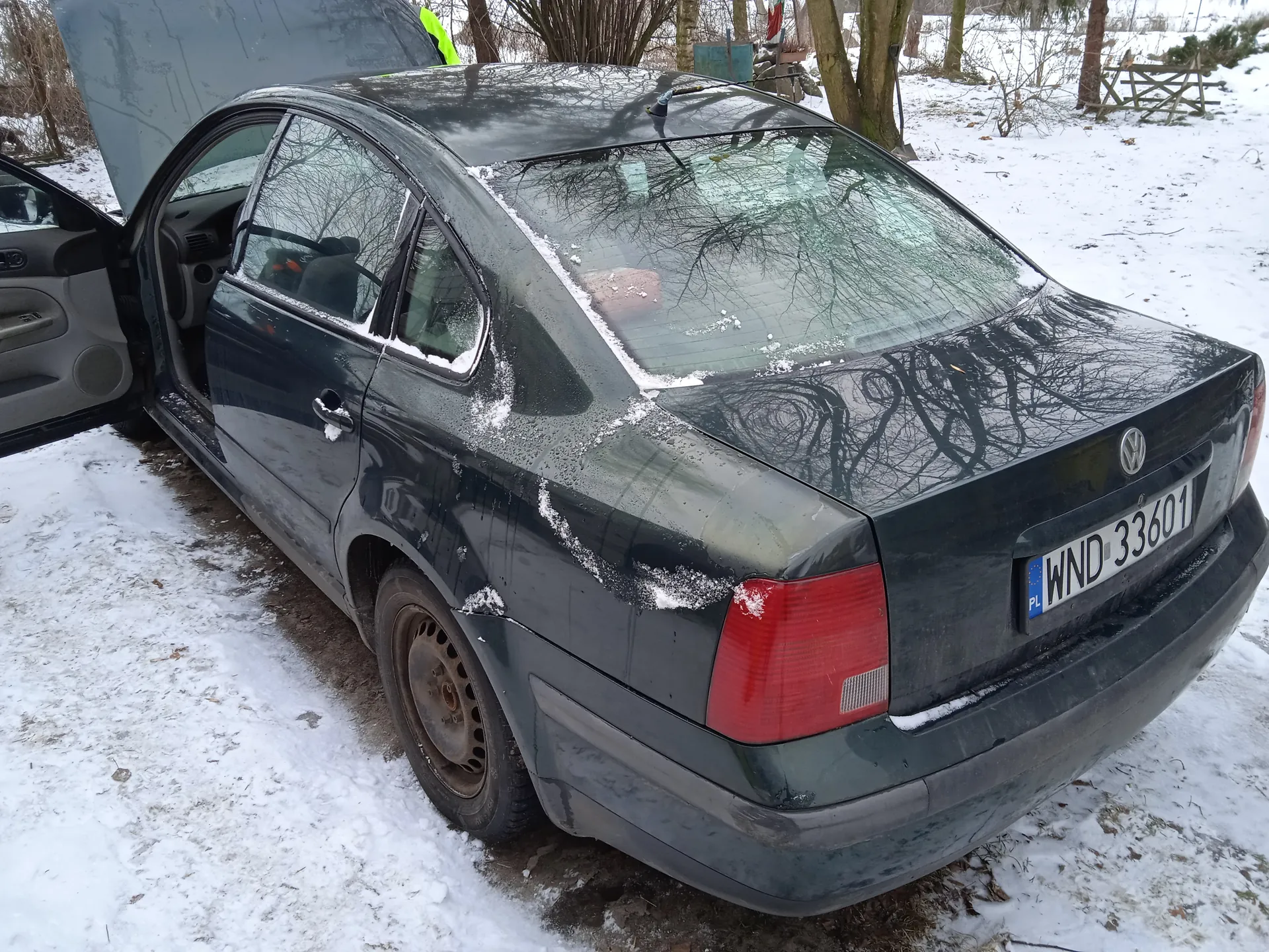 Czarny samochód osobowy marki Volkswagen z otwartym bagażnikiem i drzwiami kierowcy, zaparkowany na śnieżnej powierzchni. Na tylnej szybie i dachu widoczny śnieg. Tablica rejestracyjna z napisem WND 33601 oraz oznaczeniem PL. W tle drzewa, drewniany płot i śnieżny teren. Opona na tylnym lewym kole jest zużyta.