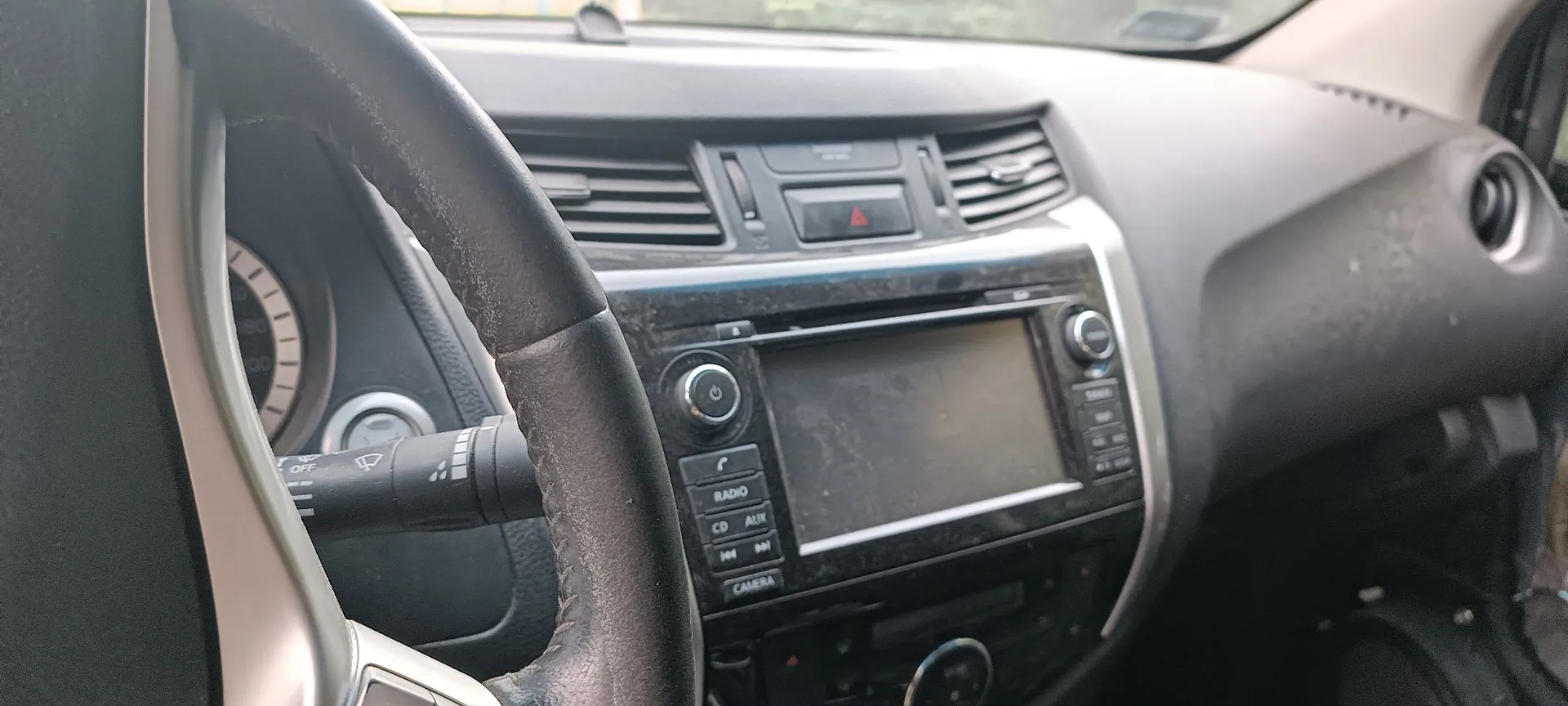 Wnętrze samochodu z perspektywy kierowcy. Widoczna jest część kierownicy, deska rozdzielcza z centralnym ekranem multimedialnym, przyciski sterujące radiem (RADIO, CD, AUX, CAMERA), nawiewy oraz czerwony przycisk alarmowy. Po lewej stronie widoczny jest fragment licznika i manetka wycieraczek.