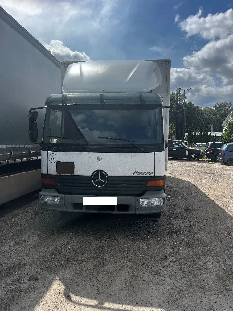 Widok z przodu białego samochodu ciężarowego marki Mercedes-Benz Atego z nadbudową. Na masce widoczne jest logo Mercedesa oraz napis 'ATEGO'. Pojazd stoi na piaszczystym placu obok innej naczepy ciężarowej. W tle widać zaparkowane samochody osobowe, drzewa oraz niebo z białymi chmurami.