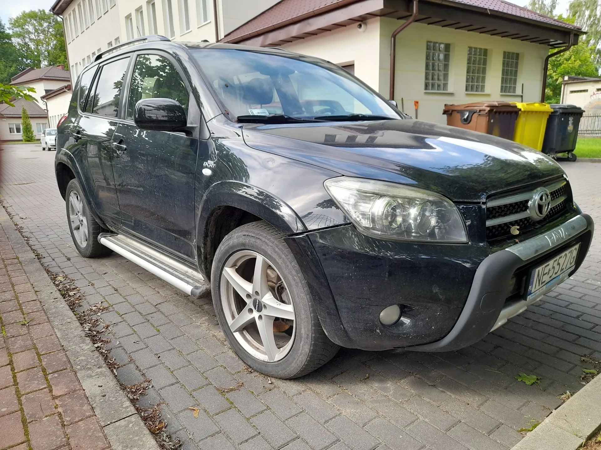 Czarny samochód terenowy marki Toyota zaparkowany na kostce brukowej przed jasnym budynkiem. Pojazd jest widoczny z przodu i z boku, posiada srebrne stopnie boczne oraz polską tablicę rejestracyjną o numerze NE 5527G. W tle widoczne są duże kontenery na odpady w kolorach brązowym i żółtym.