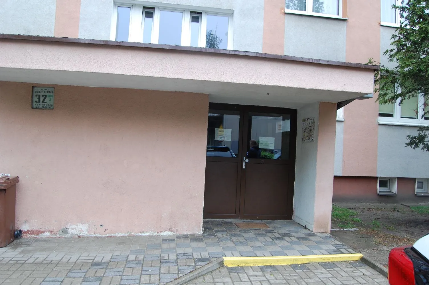 Wejście do budynku z jasnoróżową elewacją. Widoczne są brązowe, dwuskrzydłowe drzwi z przeszkleniami. Po lewej stronie drzwi, na ścianie, znajduje się zielona tabliczka z numerem 32. Przed wejściem znajduje się chodnik z szarej kostki brukowej z żółtym krawężnikiem. Nad wejściem jest betonowy daszek.