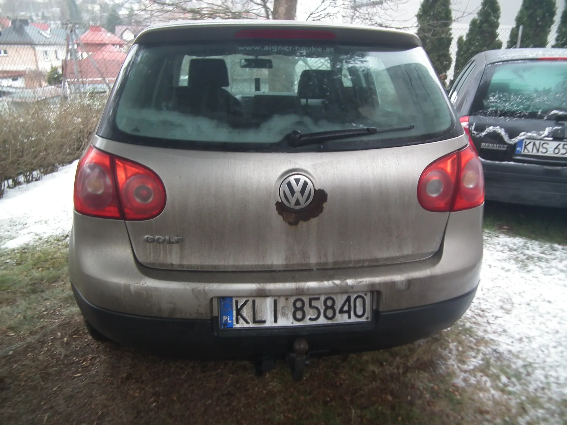 Tył srebrnego samochodu Volkswagen Golf z widocznym logo, napisem "GOLF" oraz tablicą rejestracyjną "KL 85840" z oznaczeniem "PL". Na tylnej szybie widoczny adres strony internetowej "www.aigner-hauke.at". Samochód jest zabrudzony, a wokół logo znajduje się plama rdzy. Po prawej stronie widoczny czarny samochód marki Renault z tablicą rejestracyjną "KNS 65". Tło stanowią zasneżone obszary, krzewy, drzewa, ogrodzenie i domy.